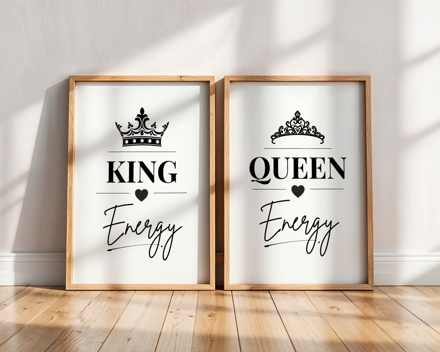 Two framed wall art prints leaning against a white wall on a wood floor. The left print says KING Energy with a crown and the word Energy, and the right print says QUEEN Energy with a tiara and the word Energy.