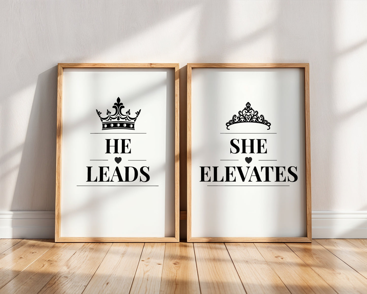 Two framed art prints leaning against a white wall on a light wood floor. The left print says 'HE LEADS' with a black crown above, and the right print says 'SHE ELEVATES' with a black tiara above.