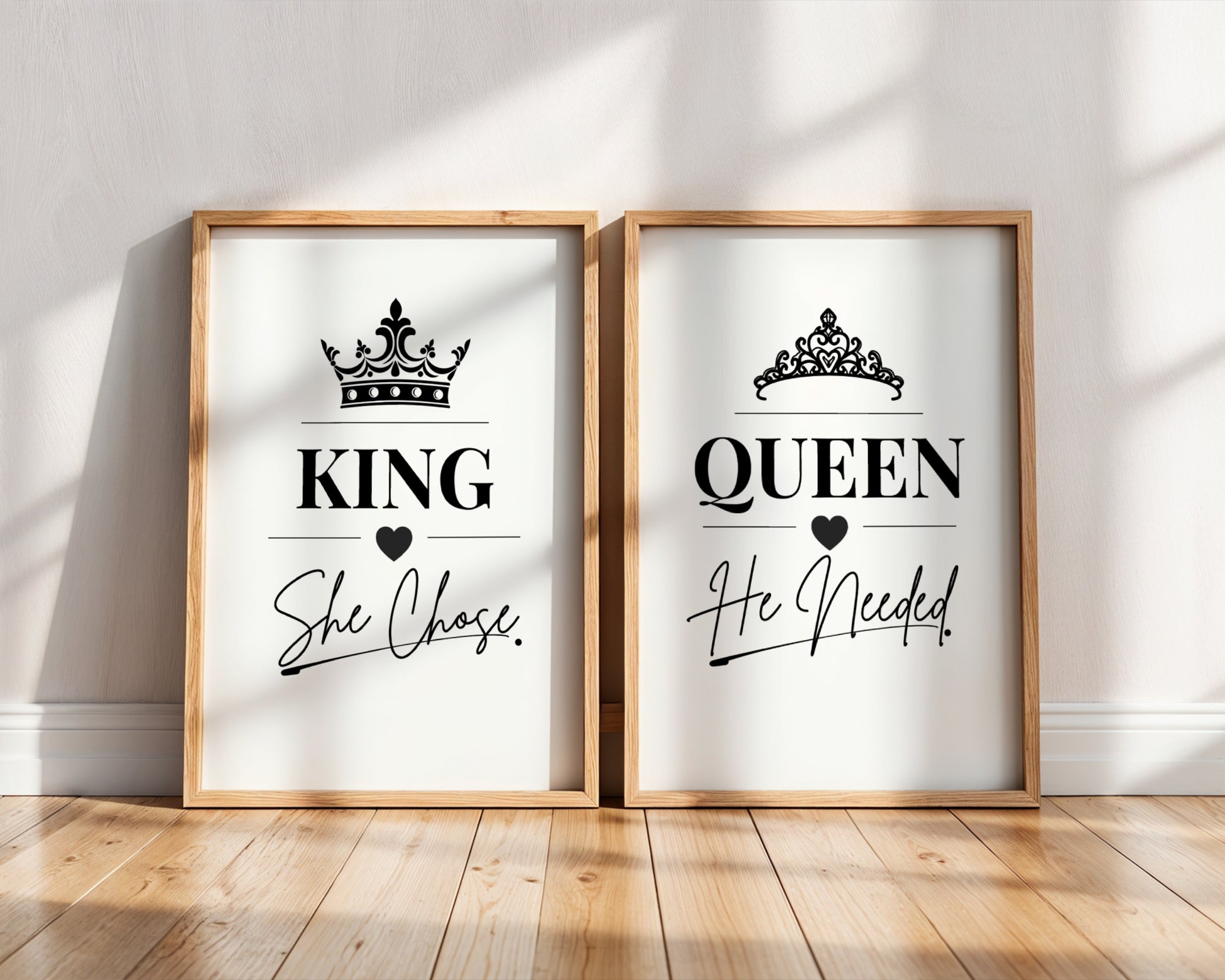 Two framed art prints leaning against a white wall on a wooden floor. The left print says 'KING She Chose' with a black crown and heart, and the right print says 'QUEEN He Needed' with a black crown and heart.