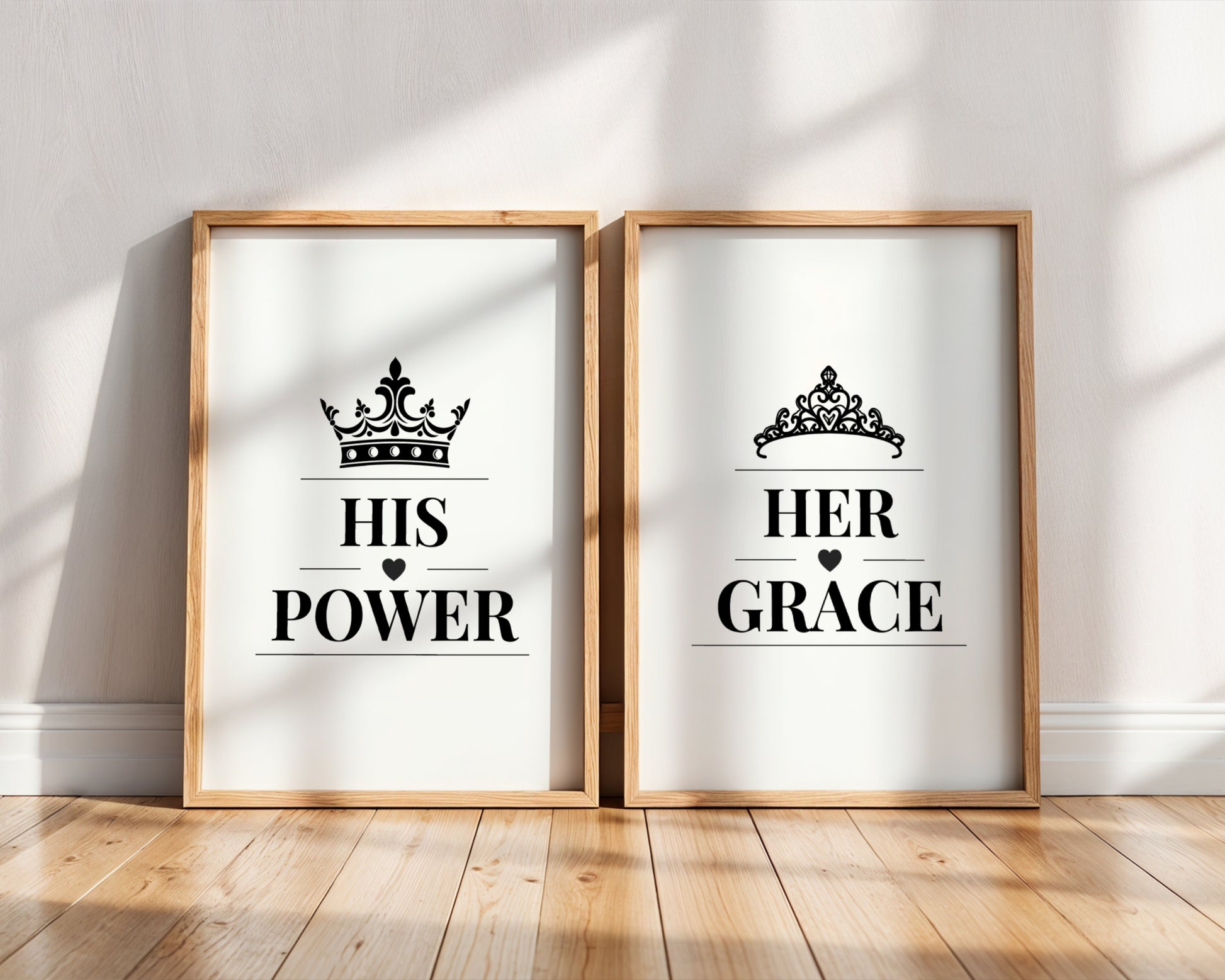 Two framed art prints leaning against a white wall on a light wood floor. The left print says 'HIS POWER' with a crown above, and the right print says 'HER GRACE' with a tiara above.