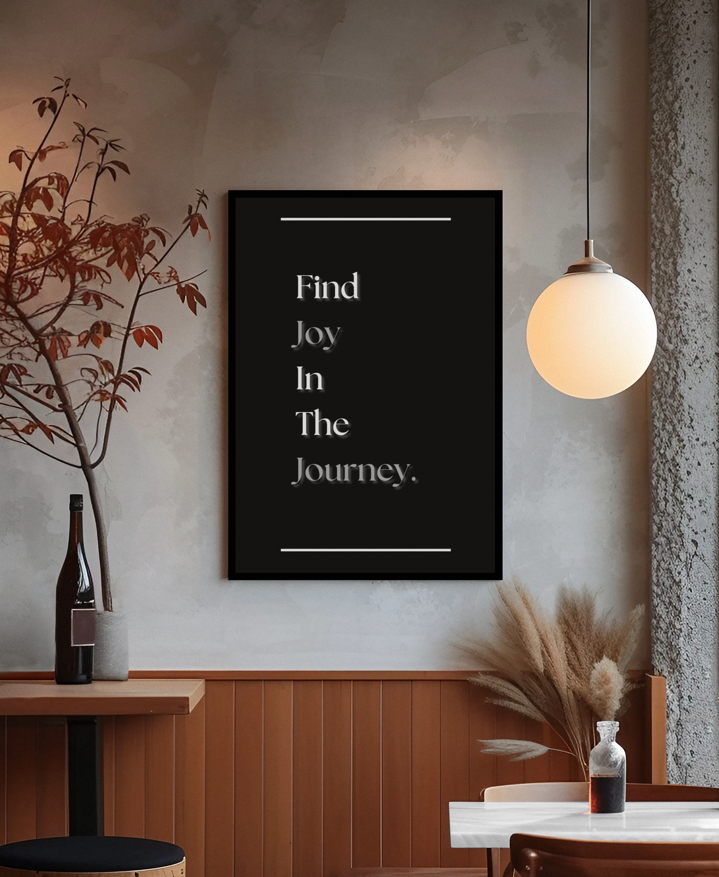 A black framed wall art print with white text reading 'Find Joy In The Journey' displayed in a modern interior setting with a warm glowing pendant light, wood-paneled wall, and decorative dried grasses.