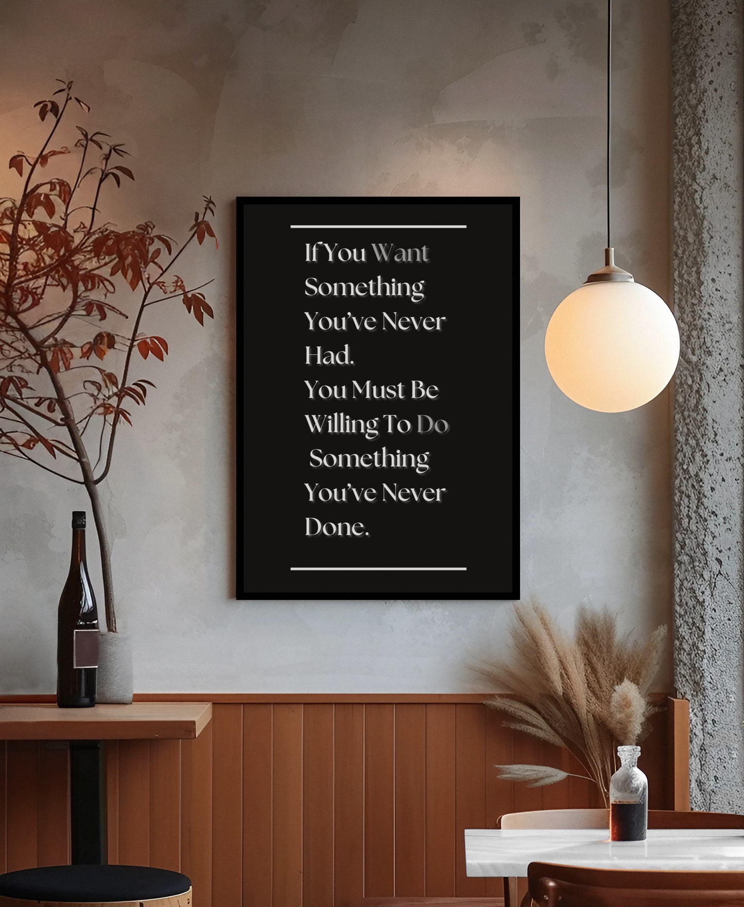 A framed motivational wall art print with white text on a black background, hanging on a textured gray wall in a modern interior setting with a hanging globe lamp, wooden paneling, and decorative dried plants.