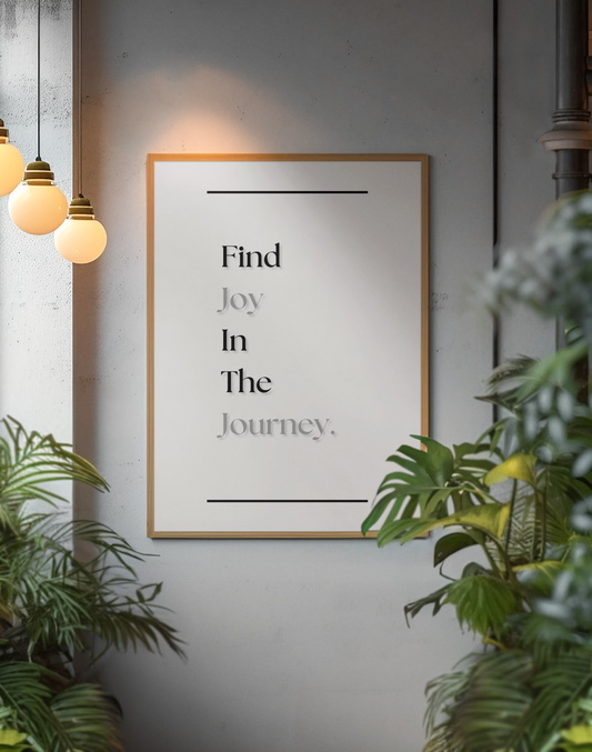 A framed typographic wall art print hanging on a gray wall, surrounded by green plants and warm hanging pendant lights. The print reads, 'Find Joy In The Journey.