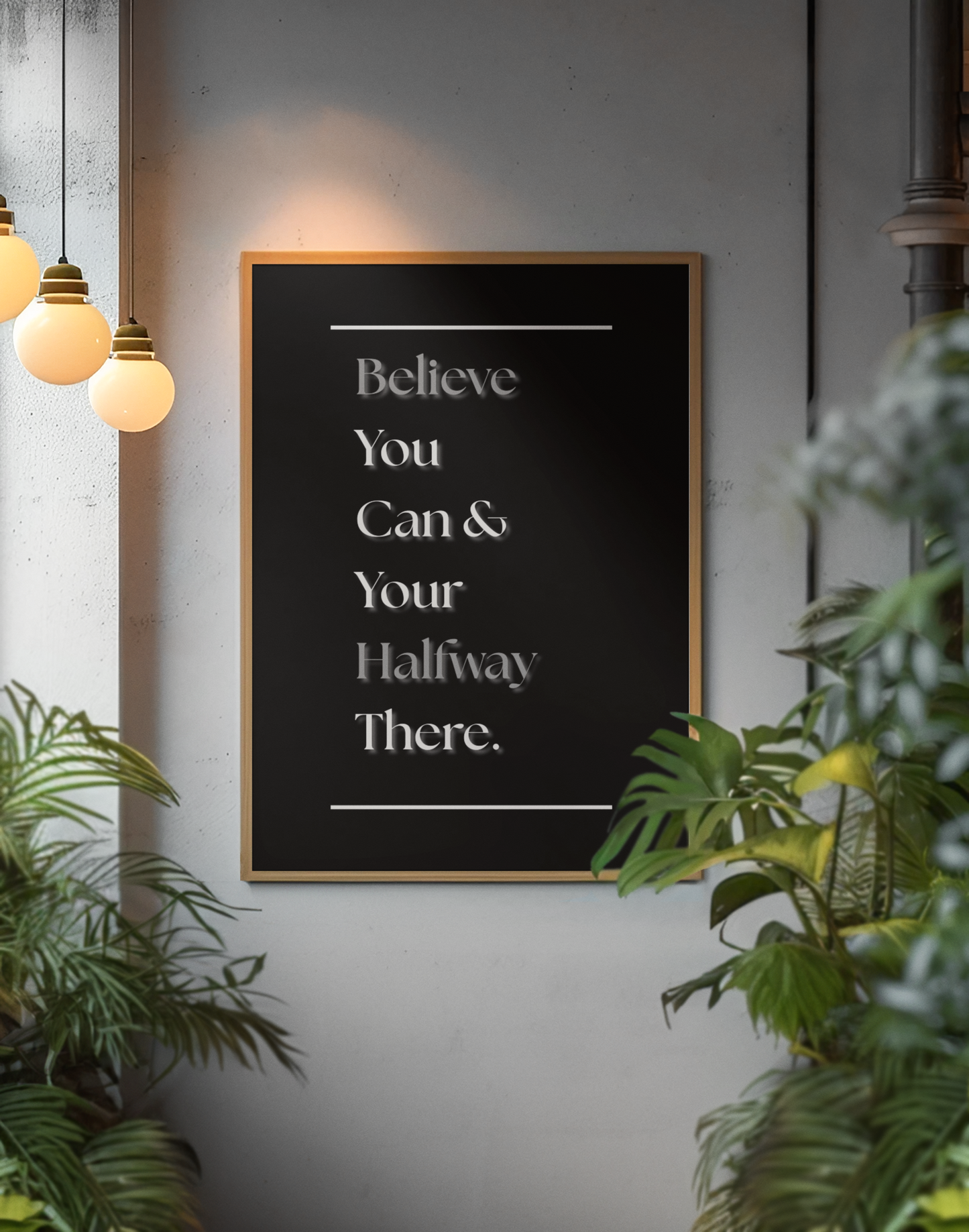 A framed motivational wall art print with the text 'Believe You Can & Your Halfway There.' in white serif lettering on a black background, hanging on a gray wall in a styled interior with hanging pendant lights and green plants.