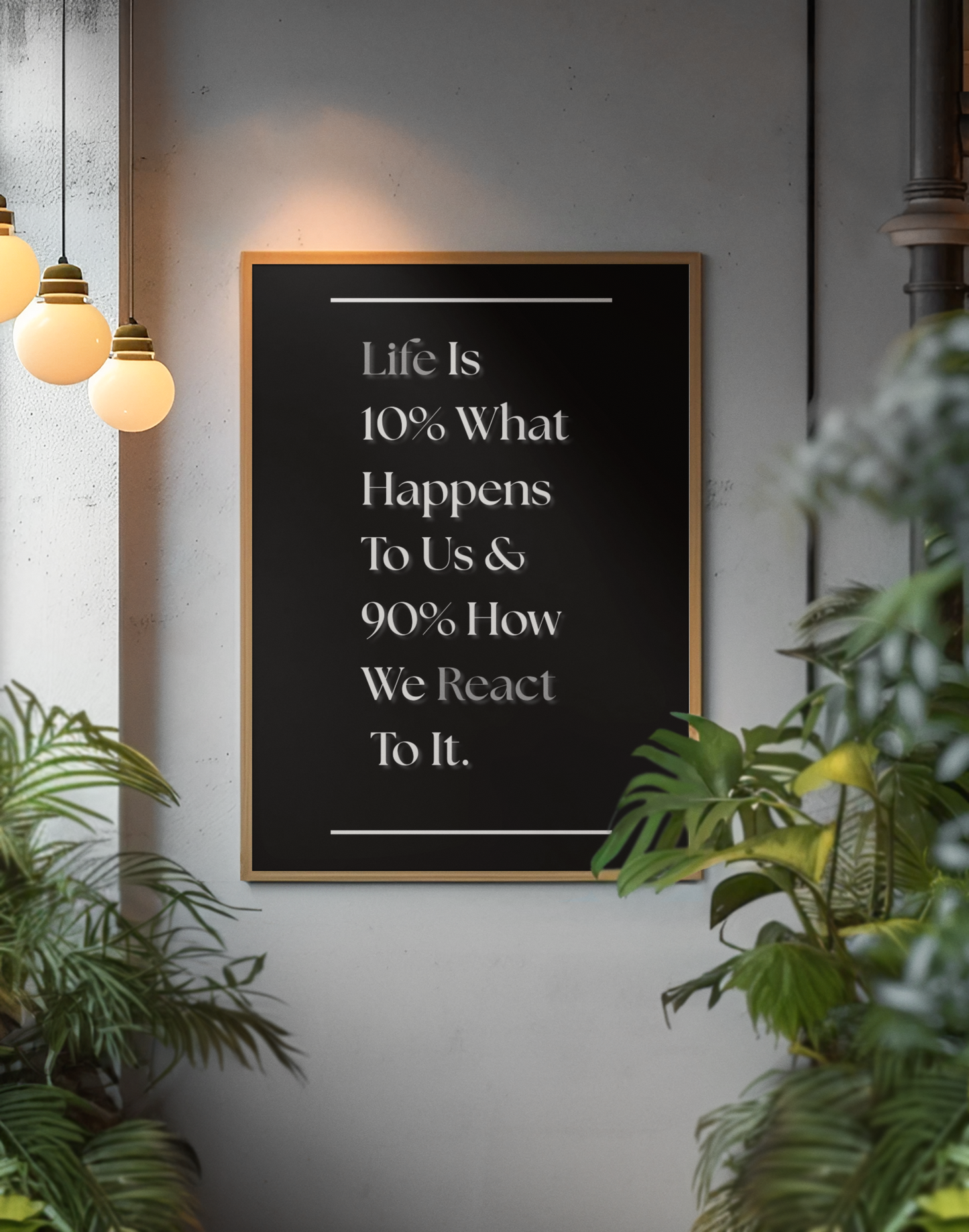 A framed motivational wall art print with the quote 'Life Is 10% What Happens To Us & 90% How We React To It.' in white serif text on a black background, hanging on a gray wall in a styled interior with hanging pendant lights and green plants.