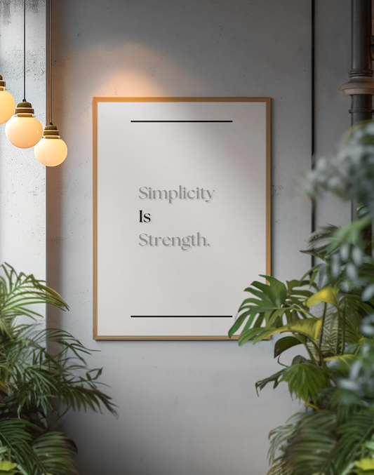 A framed wall art print with the words 'Simplicity Is Strength.' in gray serif text on a white background, hanging on a gray wall with warm pendant lights and green plants around it.