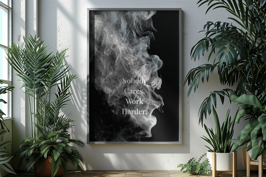 A vertical framed wall art print featuring a black background with white smoke and the text 'Nobody Cares, Work Harder.' displayed on a light wall in a styled room with potted plants.