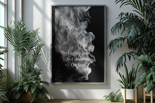 A framed vertical wall art print hanging on a light gray wall, featuring a black-and-white smoke graphic with the text 'A Future You Is Counting On You.' The room is styled with potted green plants on both sides.