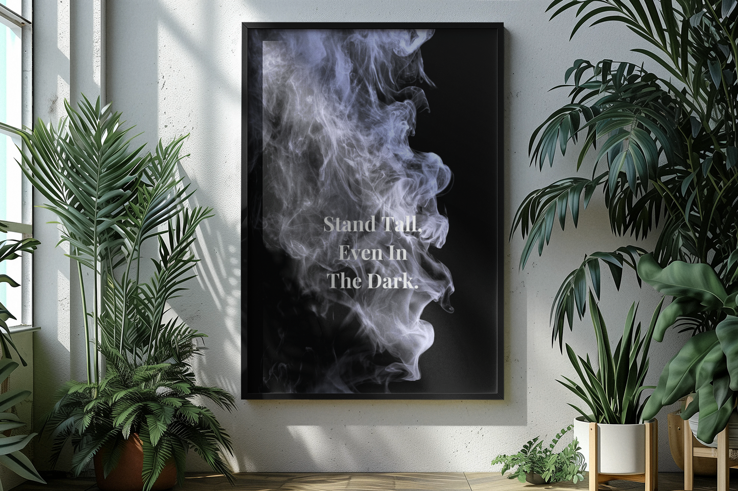 A framed vertical art print featuring a black background with a white and gray smoke illustration and the text 'Stand Tall Even In The Dark', displayed on a white wall in a styled room with potted plants.