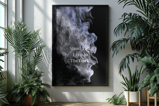 A framed vertical art print featuring a black background with a white and gray smoke illustration and the text 'Stand Tall Even In The Dark', displayed on a white wall in a styled room with potted plants.