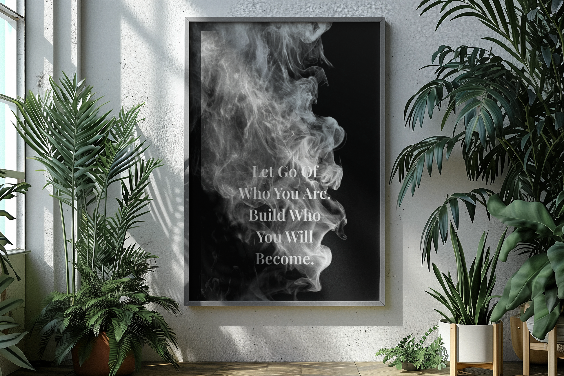 A vertical framed wall art print with a black background, white smoke graphic, and the text 'Let Go Of Who You Are, Build Who You Will Become,' displayed on a light wall between large green potted plants.