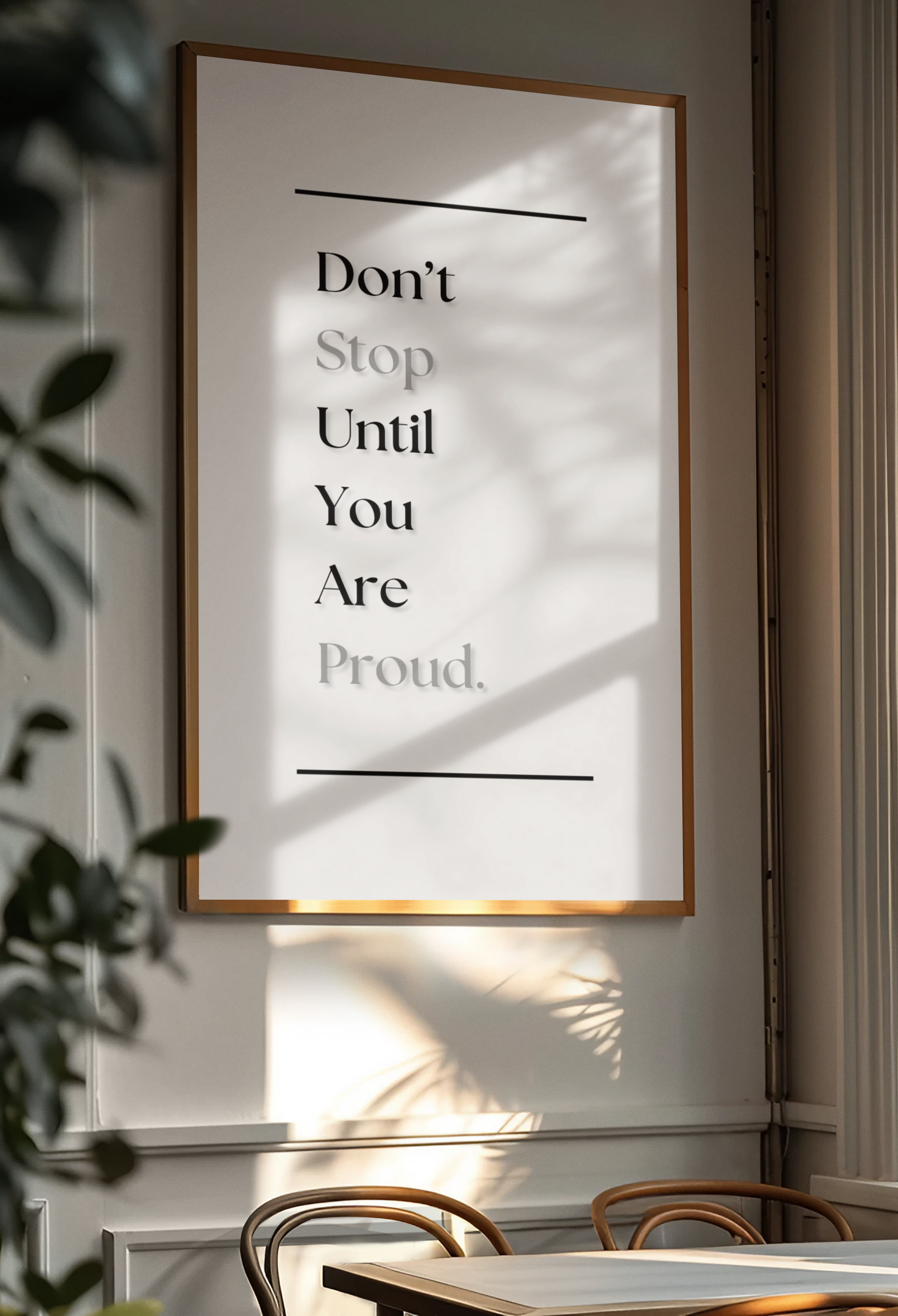 A framed typographic wall art print hanging on a light wall in a real interior setting, with sunlight and leaf shadows cast across the print. The print reads, 'Don't Stop Until You Are Proud.