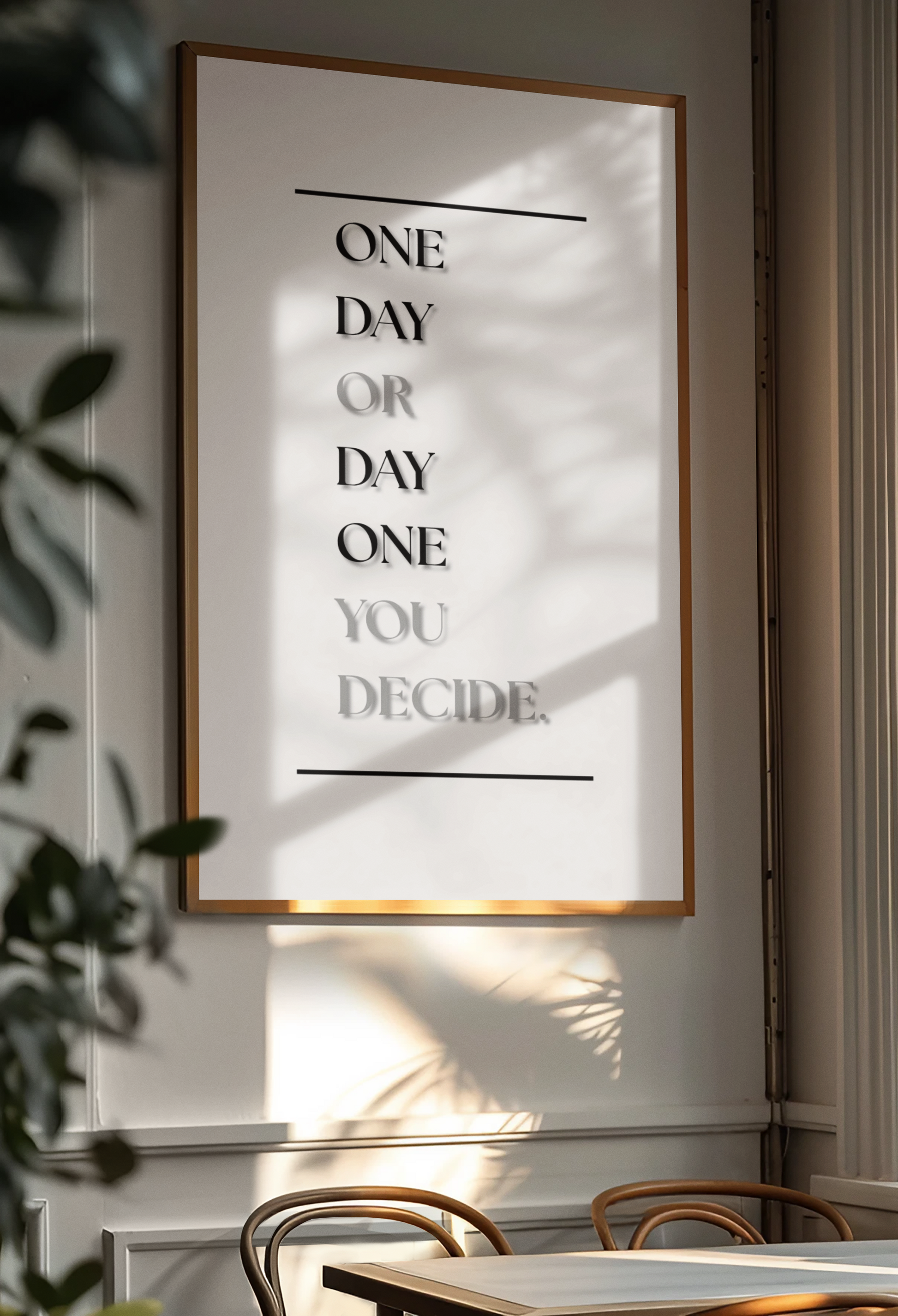 A framed typographic wall art print hanging on a light wall in a real interior setting, with sunlight and leaf shadows cast across it. The print contains only black text on a white background: 'One Day Or Day One, You Decide.'