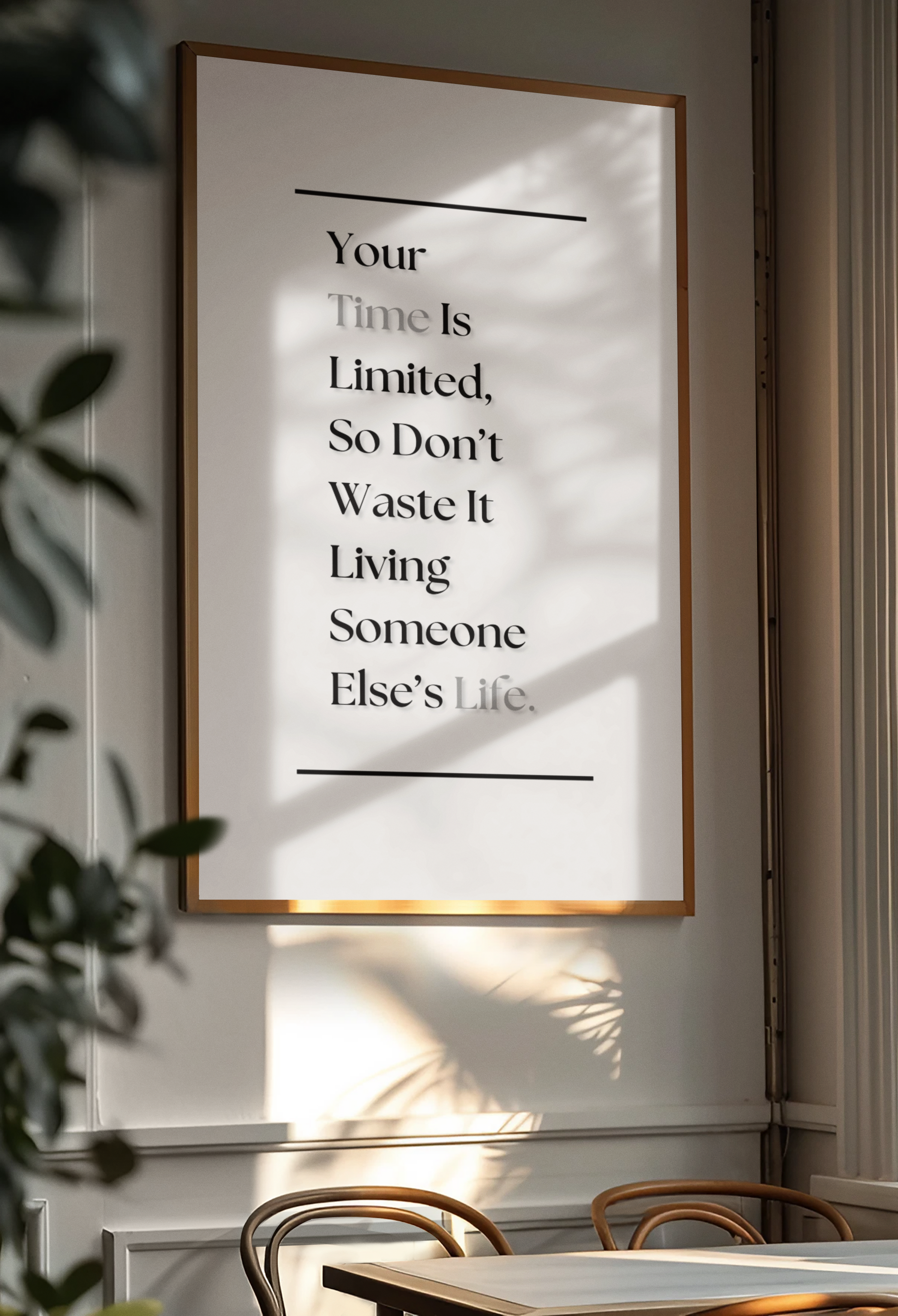 A framed typographic wall art print hanging on a light wall in a real interior setting, with sunlight and leaf shadows cast across the print. The print contains a motivational quote in black serif text on a white background with a thin warm-toned frame.