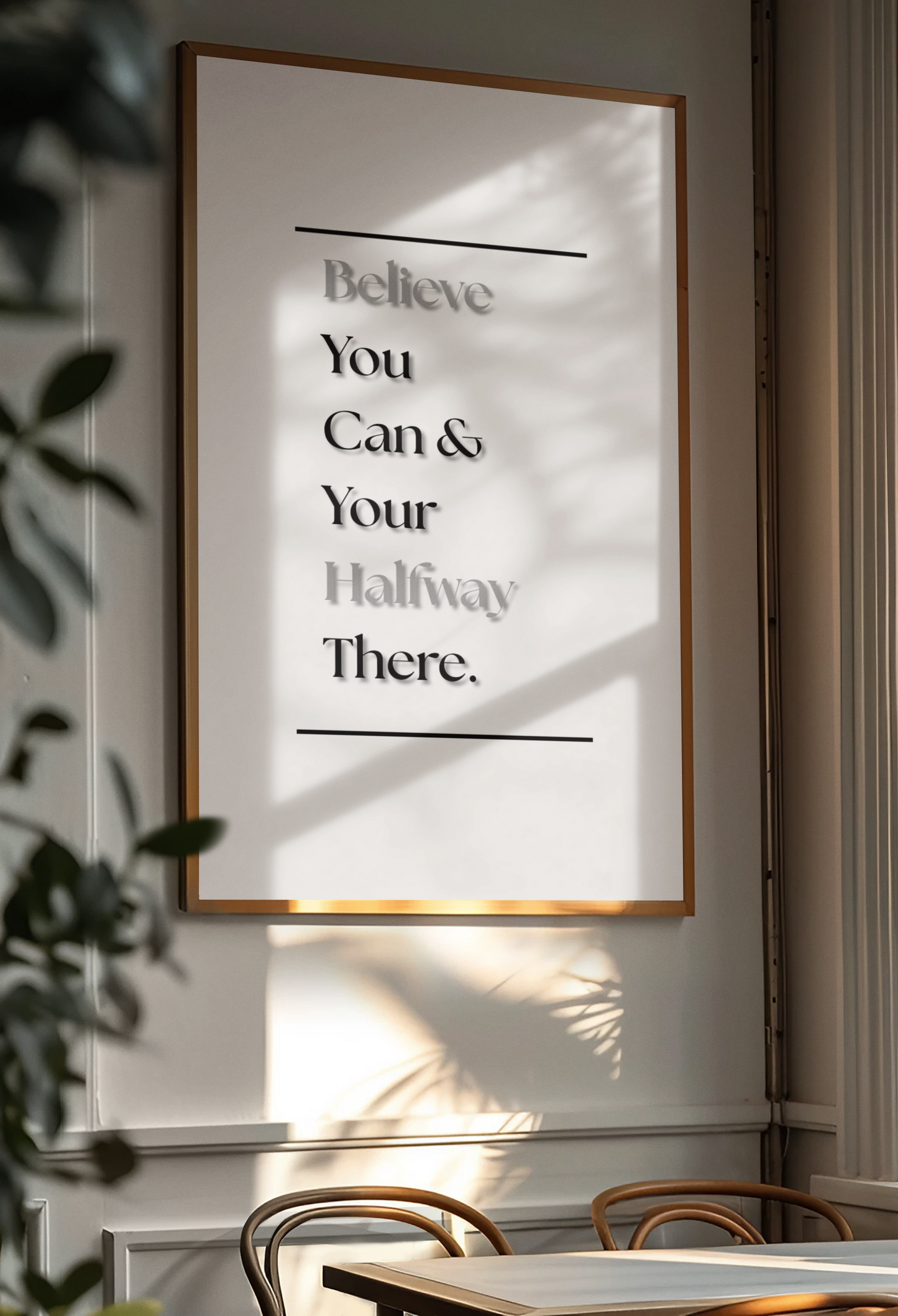 A framed motivational wall art print with the text 'Believe You Can & Your Hallway There.' hanging on a light gray wall in a dining area, with warm sunlight and leaf shadows cast across it.