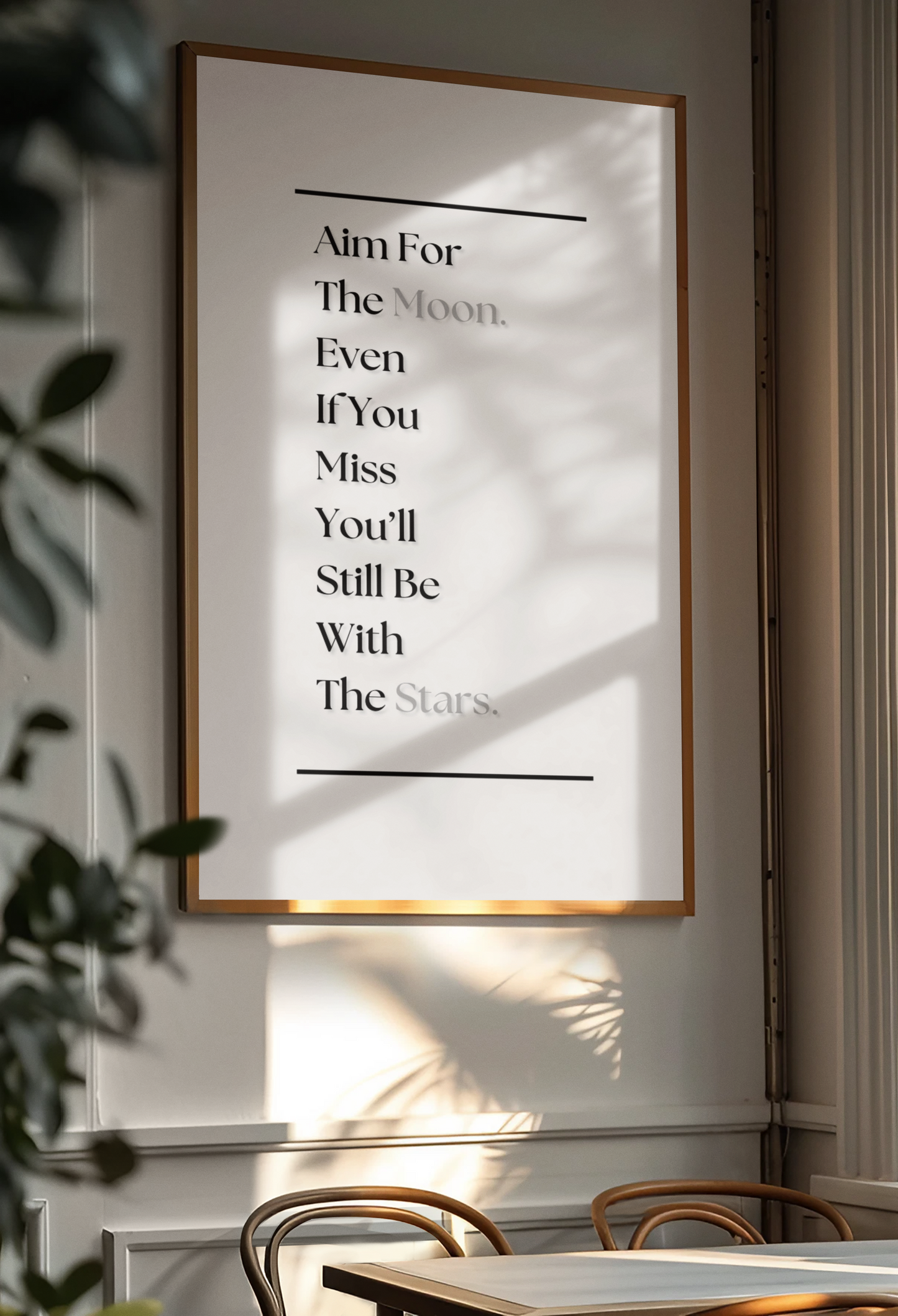 A framed wall art print with a minimalist quote hangs on a light gray wall in a real interior setting with sunlight, leaf shadows, and part of a dining table and chairs visible.