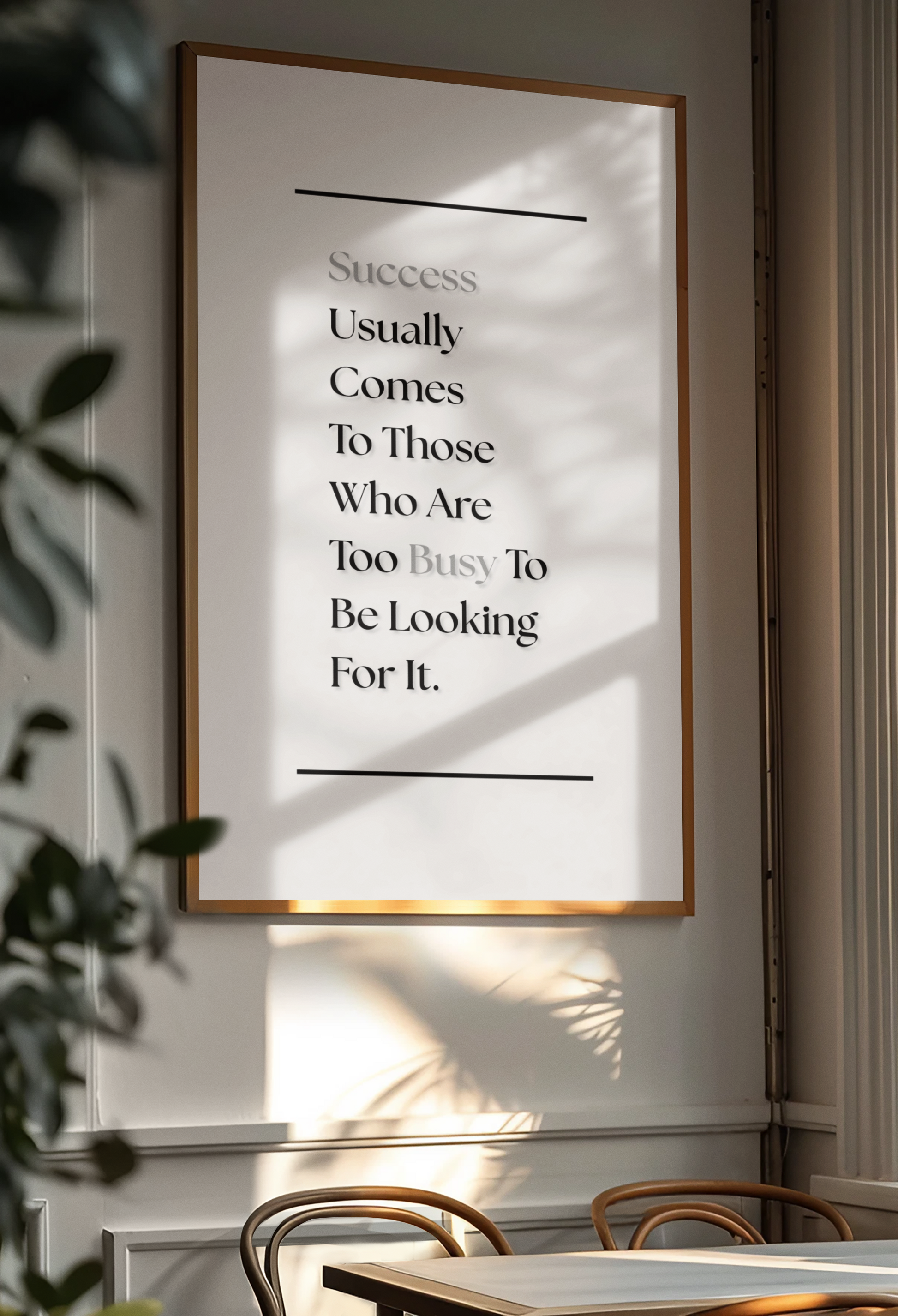 A framed motivational wall art print with the quote 'Success Usually Comes To Those Who Are Too Busy To Be Looking For It.' displayed on a light gray wall in a dining room setting with warm sunlight and leaf shadows.