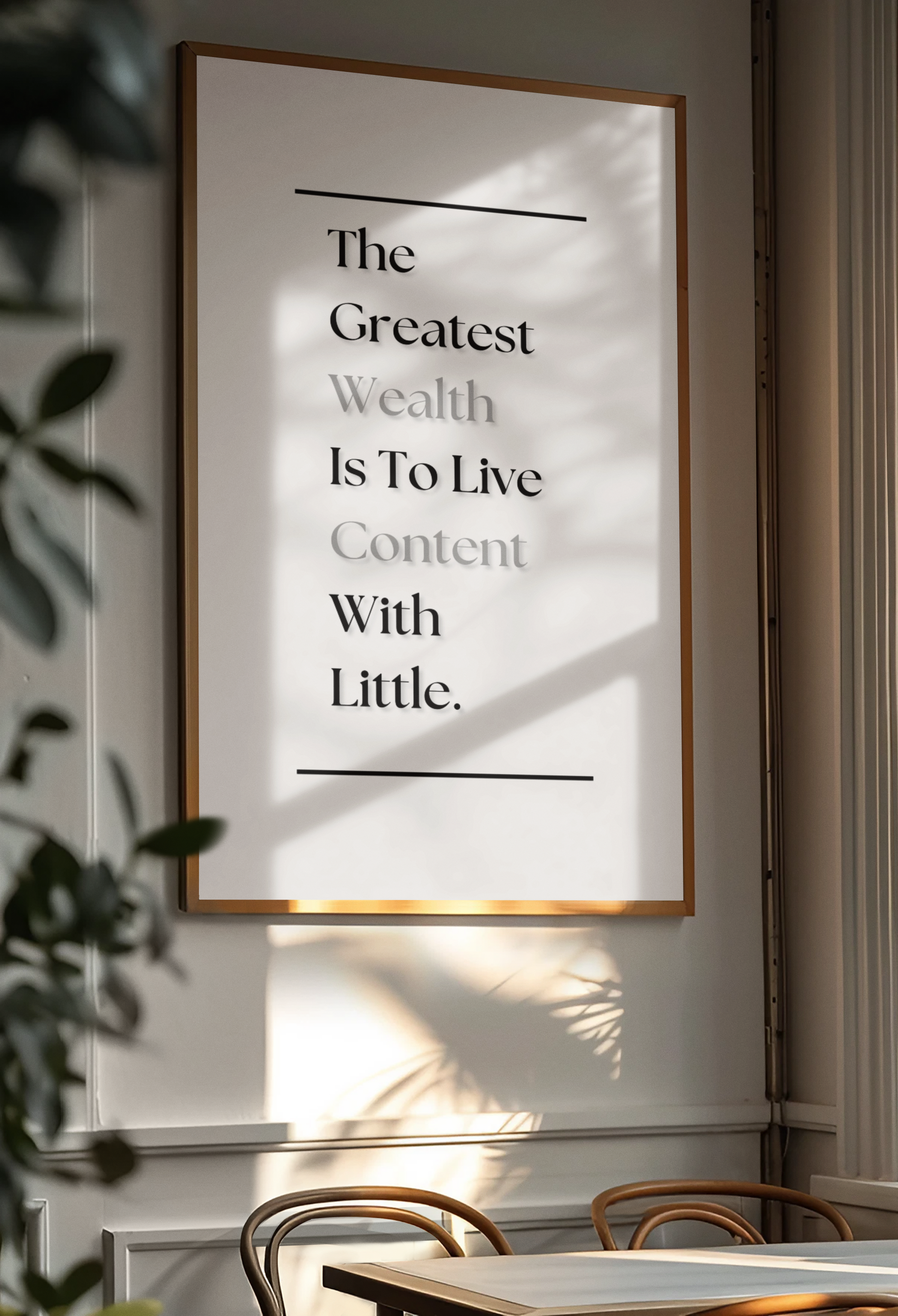 A framed typographic wall art print hanging on a light gray wall in a real interior setting, with sunlight casting shadows across the print and table chairs visible below. The print contains the quote, 'The Greatest Wealth Is To Live Content With Little'.