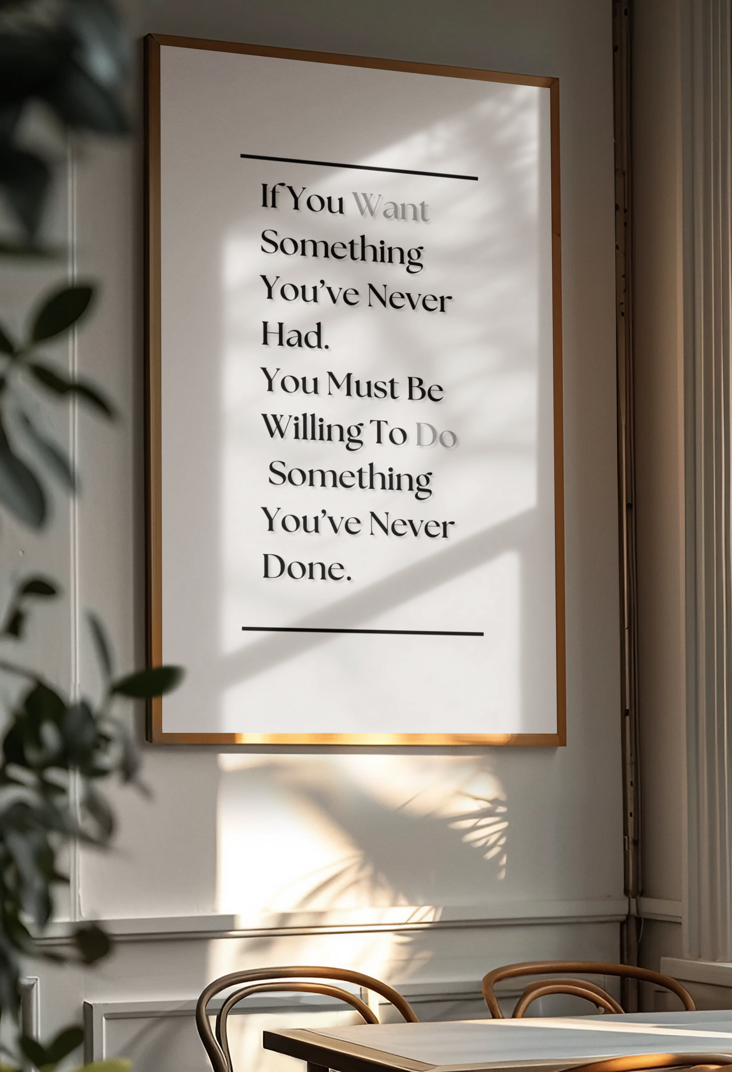 A framed motivational poster with a gold-toned frame hanging on a light gray wall. The poster features a black serif quote on a white background, styled in a dining room setting with warm sunlight and leaf shadows cast across it.