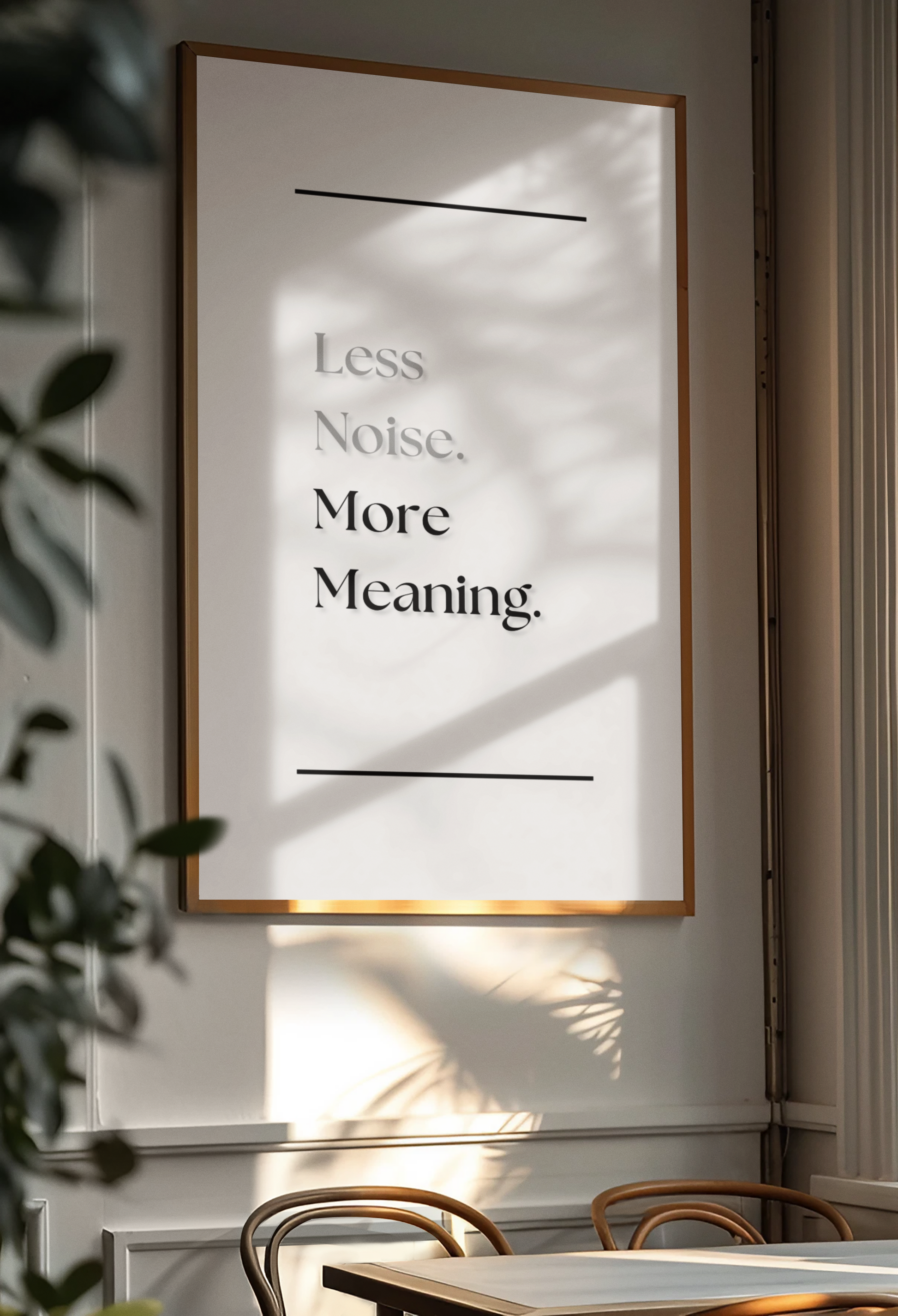 A framed motivational wall art print with the text 'Less Noise. More Meaning.' in black serif font on a white background, hanging on a light wall in a real interior setting with sunlight, shadows, and part of a table and chairs visible.