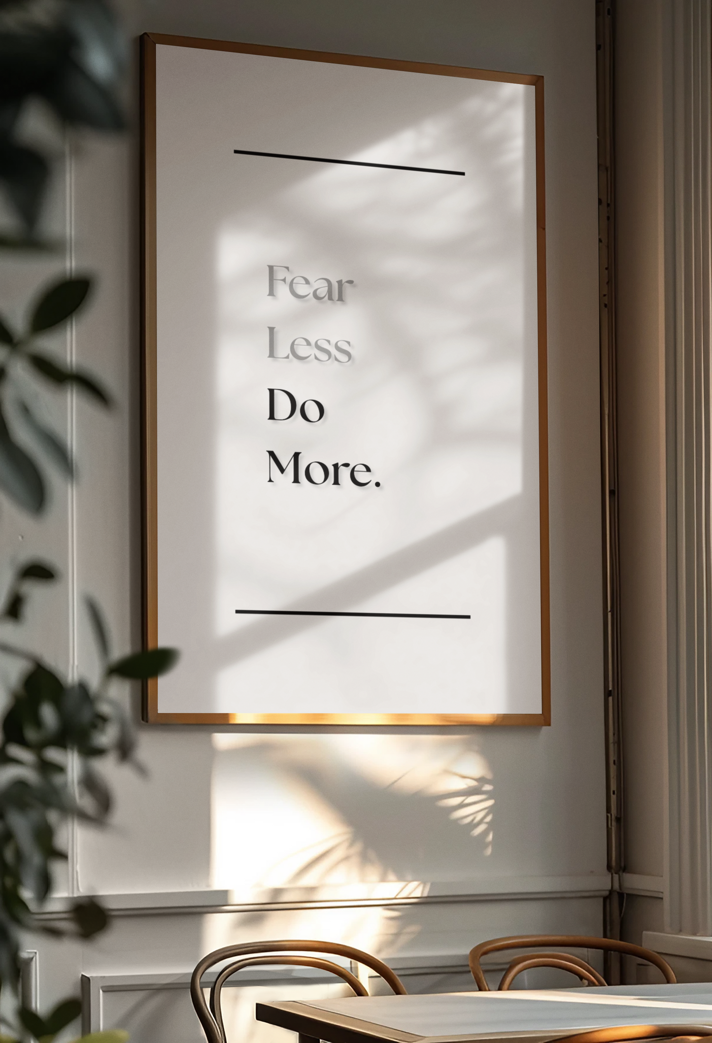 A framed minimalist art print with the text 'Fear Less, Do More.' hangs on a light gray wall in a dining room setting, with warm sunlight and leaf shadows cast across the print.
