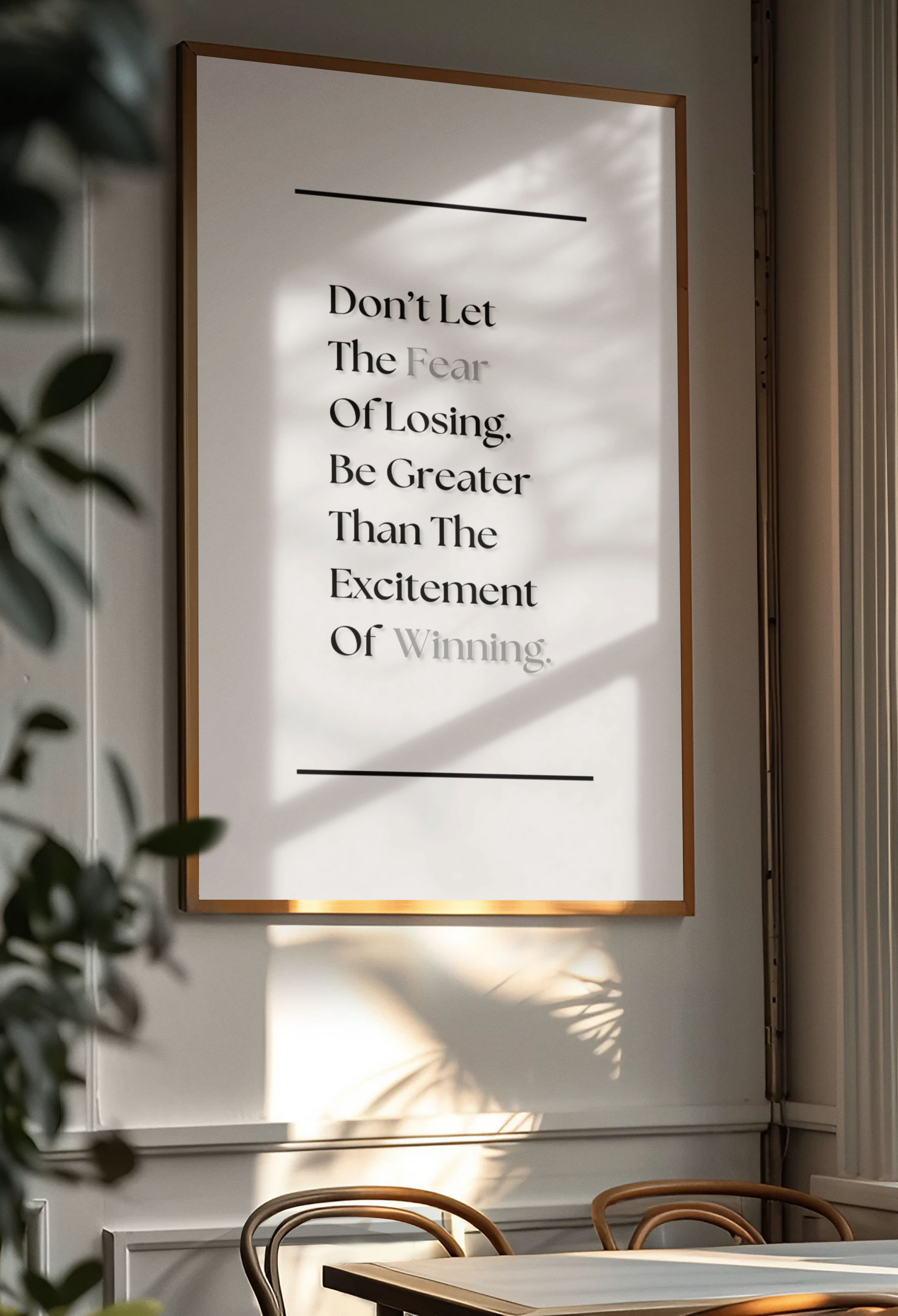 A framed motivational wall art print with the quote 'Don't Let The Fear Of Losing Be Greater Than The Excitement Of Winning' in black serif text on a white background, hanging on a light gray wall in a styled interior with soft leaf shadows.
