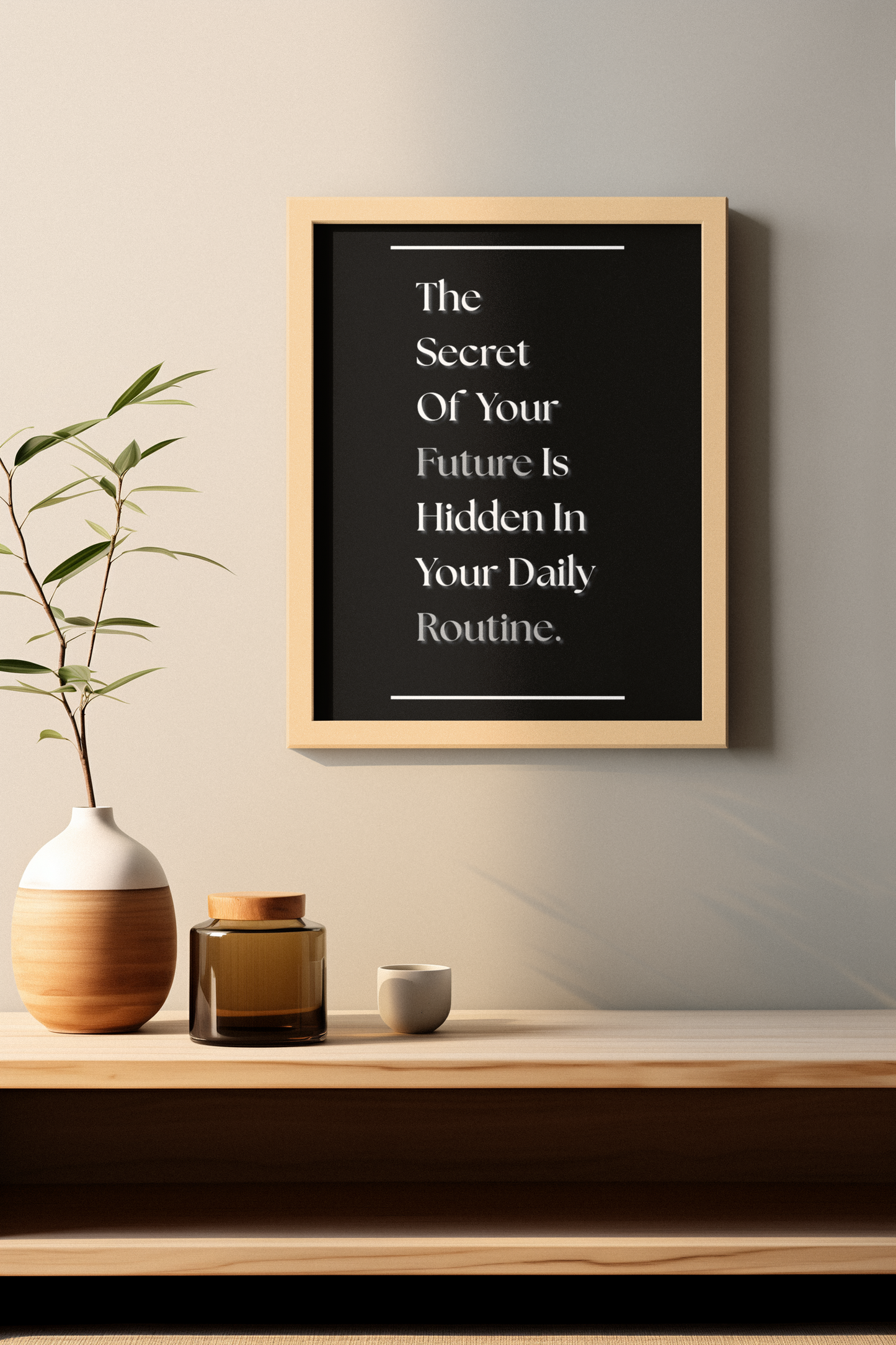 A framed typographic wall art print hangs on a light beige wall above a wooden shelf. The print has a black background with white text reading, 'The Secret Of Your Future Is Hidden In Your Daily Routine'.