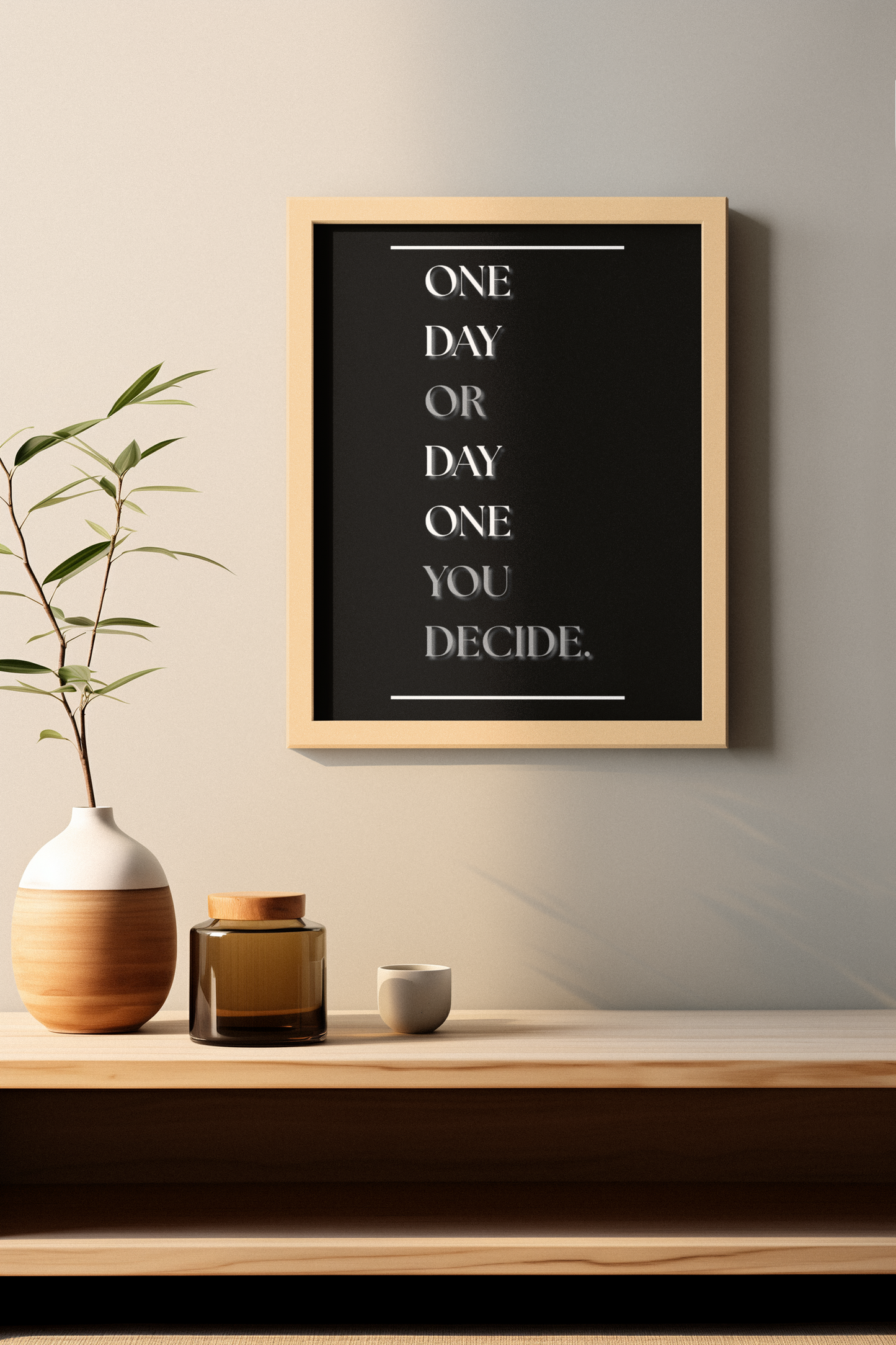 A framed typographic wall art print hangs on a beige wall above a wooden shelf. The print has a black background with white text reading, 'One Day OR Day One, You Decide.'