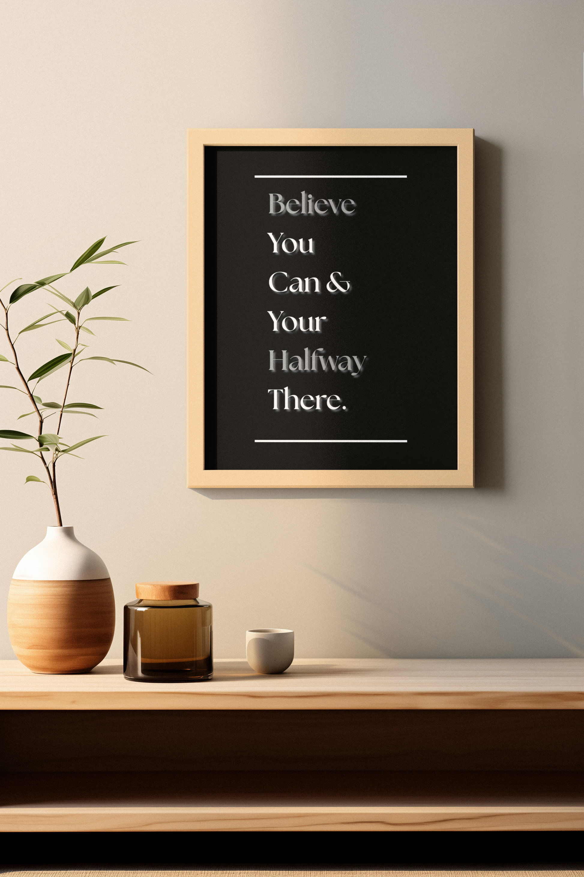 A framed motivational wall art print with the quote 'Believe You Can & Your Halfway There', in white text on a black background, styled in a minimalist interior setting above a wooden shelf with a vase and small decor items.