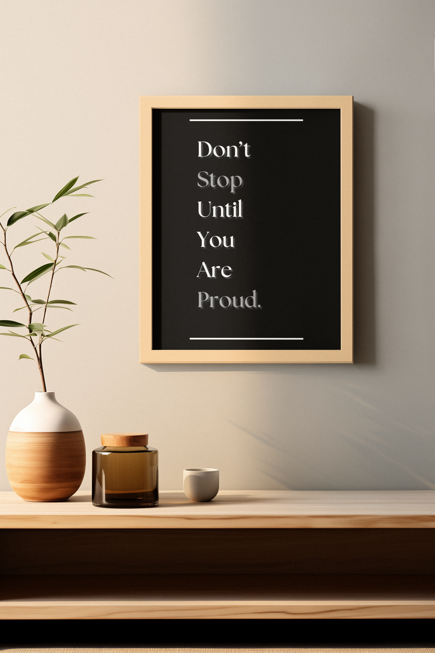 A framed motivational wall art print with the text 'Don't Stop Until You Are Proud.' in white on a black background, hanging on a beige wall above a wooden shelf with decorative ceramic items and a plant.