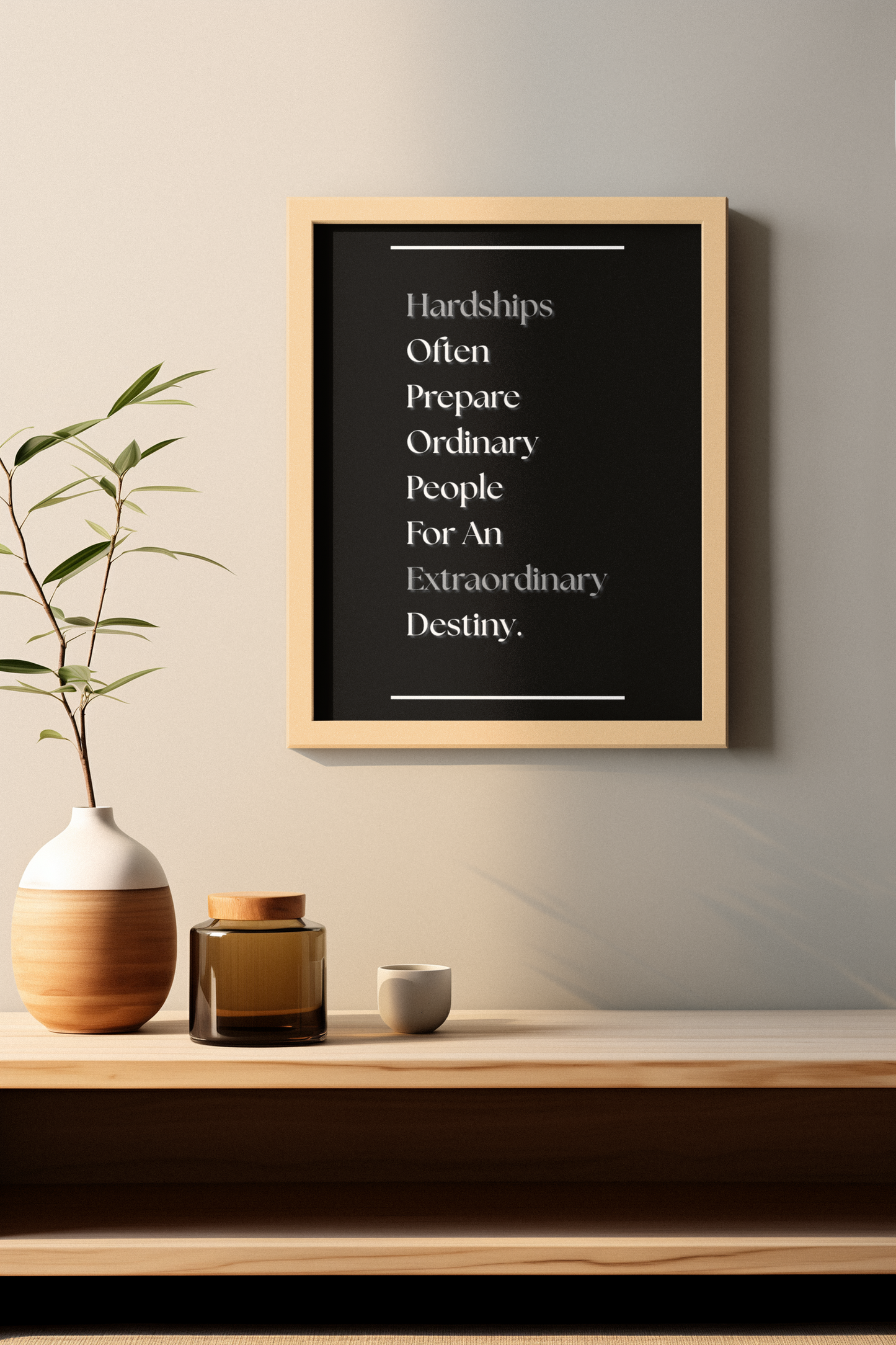 A framed motivational wall art print with the quote 'Hardships Often Prepare Ordinary People For An Extraordinary Destiny.' displayed on a light wood shelf above a ceramic vase and small jars against a neutral beige wall.