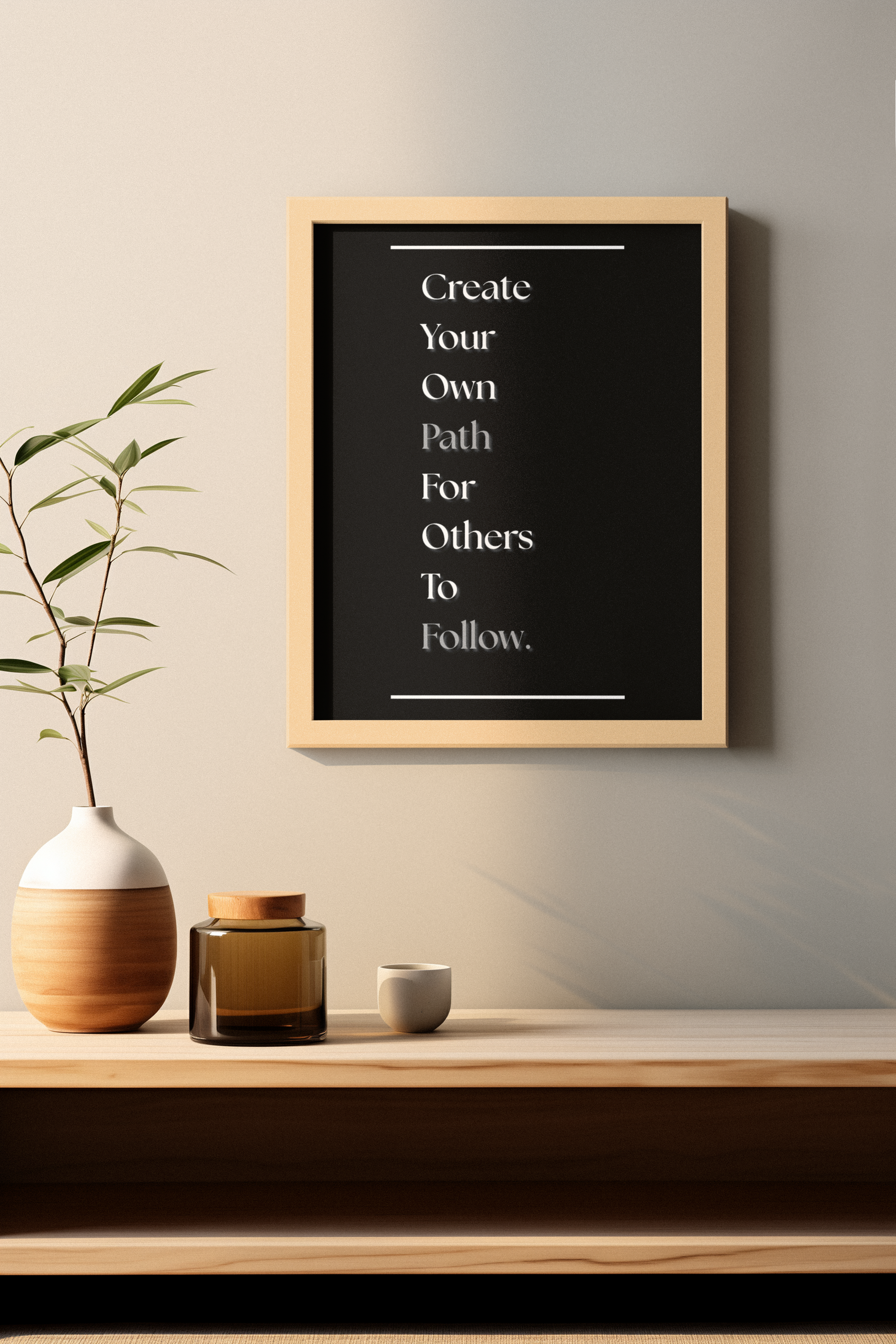 A framed wall art print with the quote 'Create Your Own Path For Others To Follow.' in white text on a black background, hanging above a wooden shelf with a vase, small jar, and cup in a minimalist interior setting.