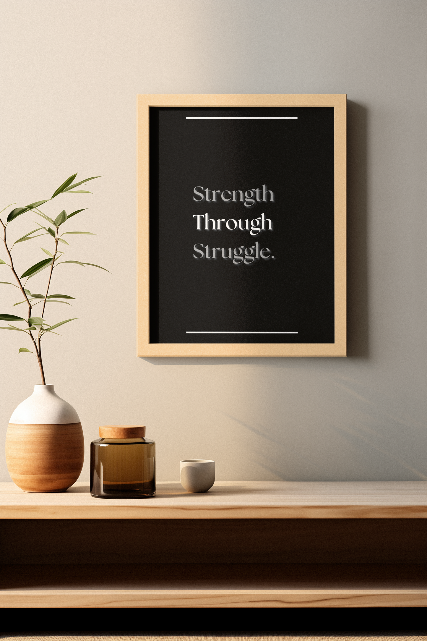 A framed typographic wall art print reading 'Strength Through Struggle' in white serif text on a black background, hanging above a wooden console table with a vase, amber jar, and small cup in a styled interior setting.