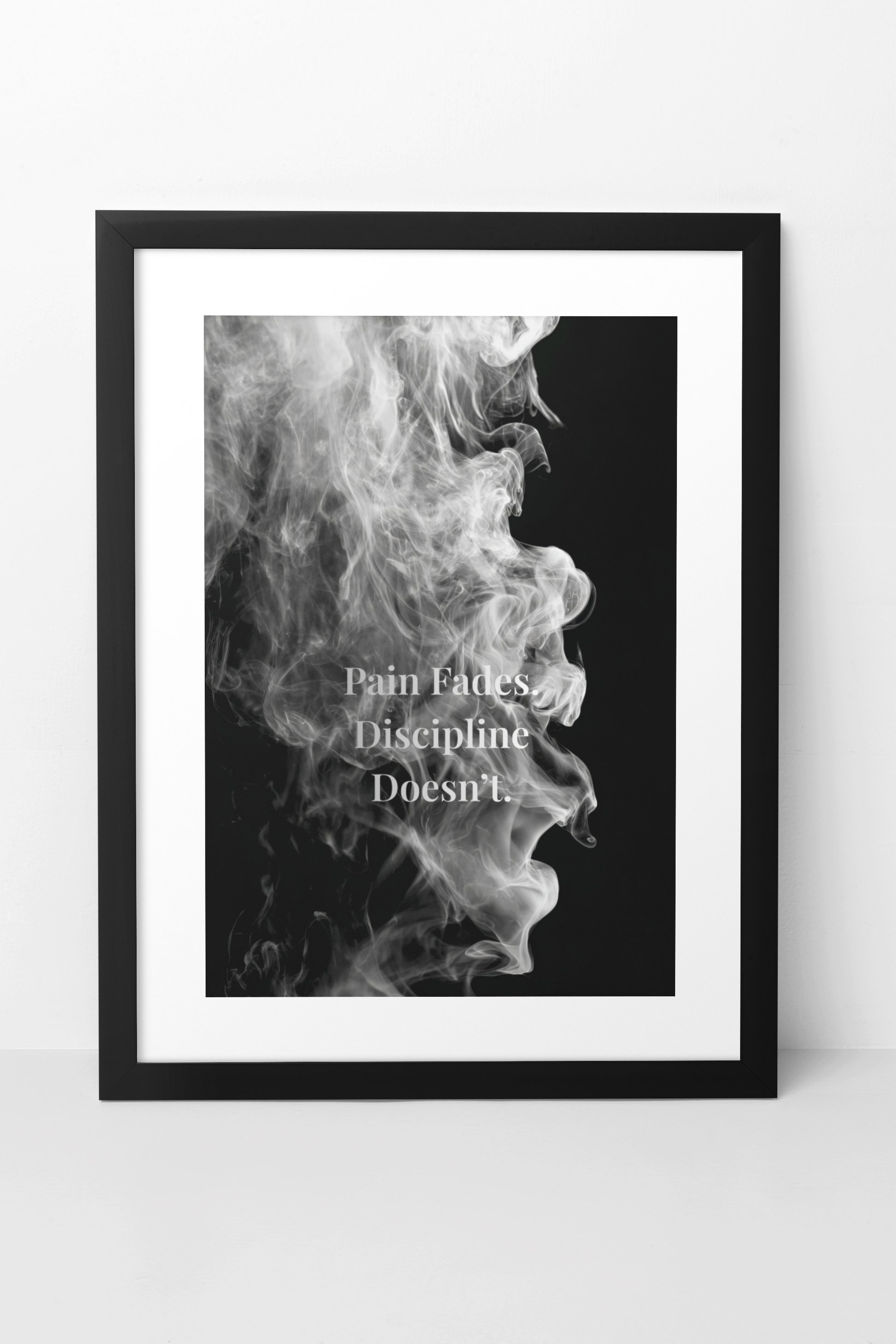 A framed black and white art print featuring a profile of smoke against a black background with the quote 'Pain Fades Discipline Doesn't' in white text, shown in a black frame on a light gray wall.