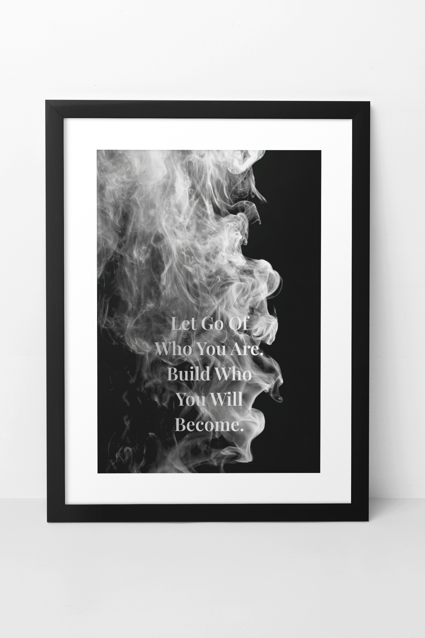 A framed black and white motivational poster featuring a smoky graphic and the quote 'Let Go Of Who You Are, Build Who You Will, Become.' The print is shown in a black frame with a white mat against a light gray wall.