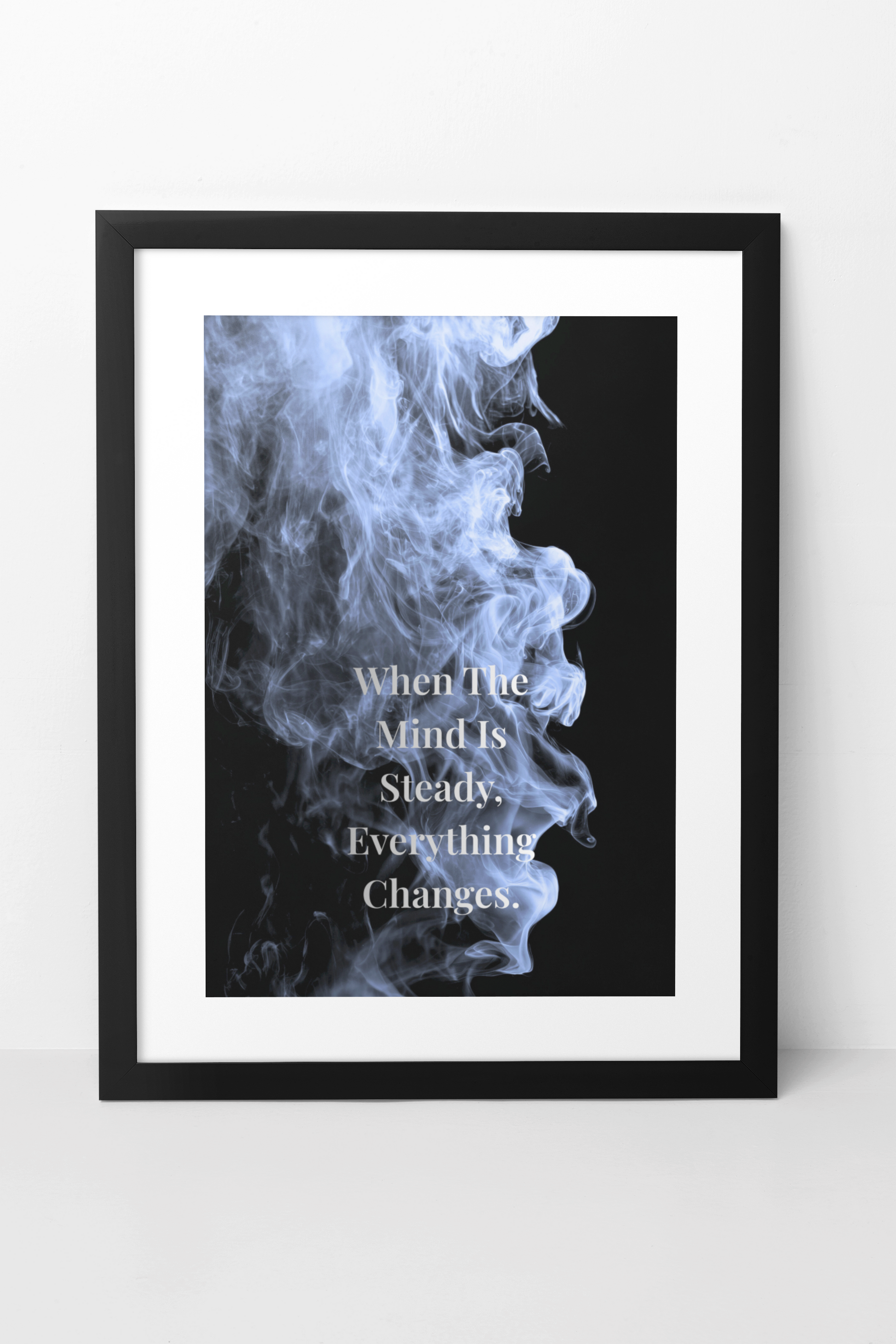 A framed art print featuring a black background with a blue-gray smoke profile and white text reading, 'When The Mind Is Steady, Everything Changes.' The print is matted in white and enclosed in a slim black frame, shown against a light gray wall and floor.