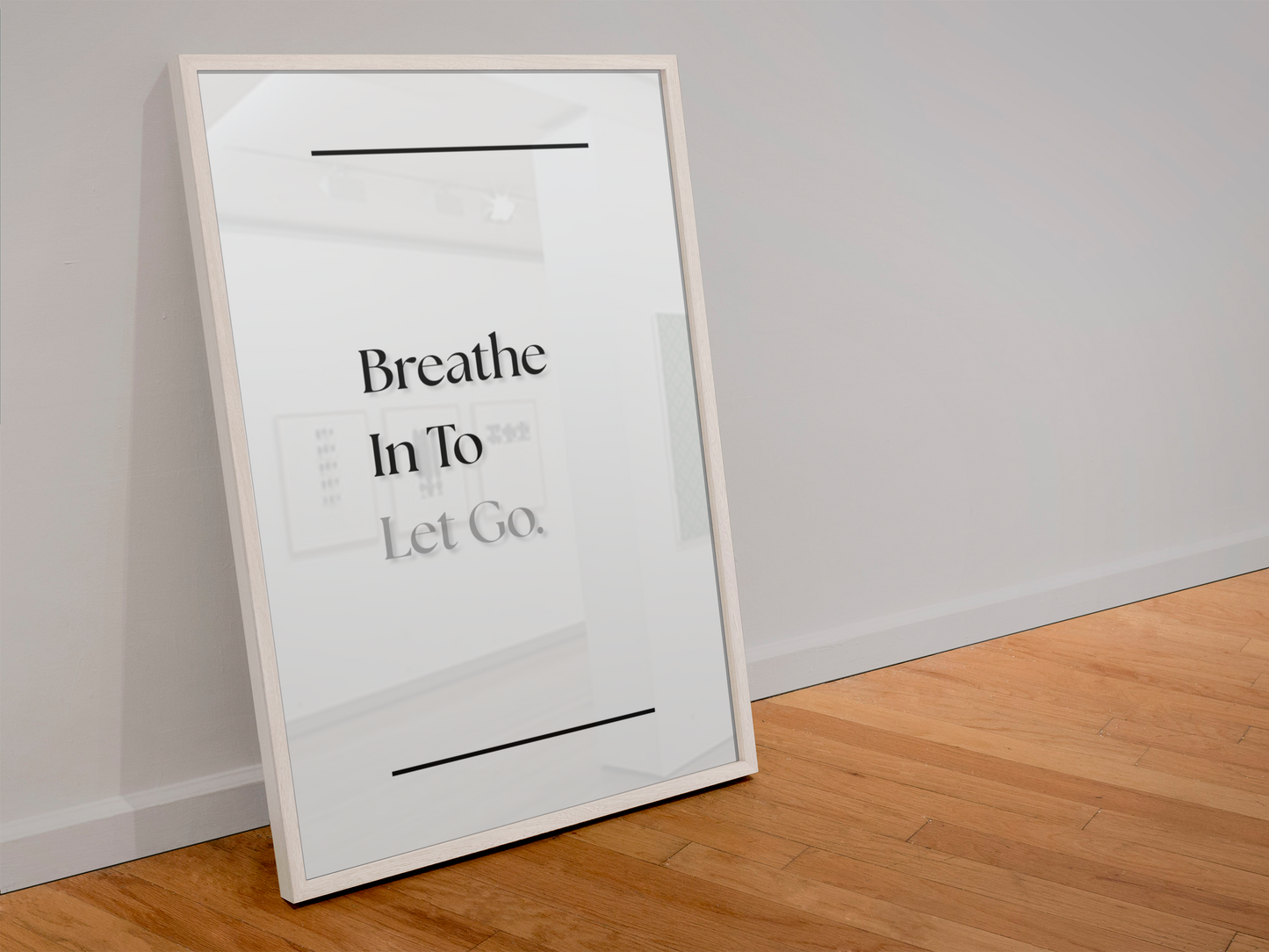A framed minimalist art print leaning against a light gray wall on a light wood floor. The print features the text 'Breathe In To Let Go'.