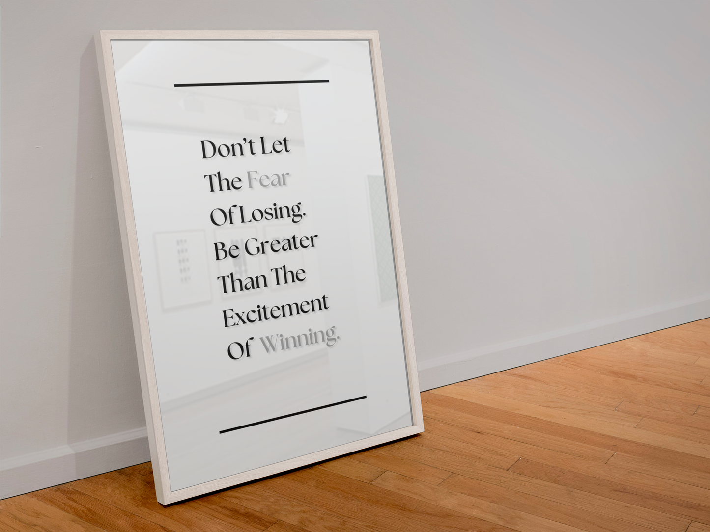 A framed motivational poster leaning against a white wall on a light wood floor. The poster features a white background with black text that reads, 'Don't Let The Fear Of Losing Be Greater Than The Excitement Of Winning.