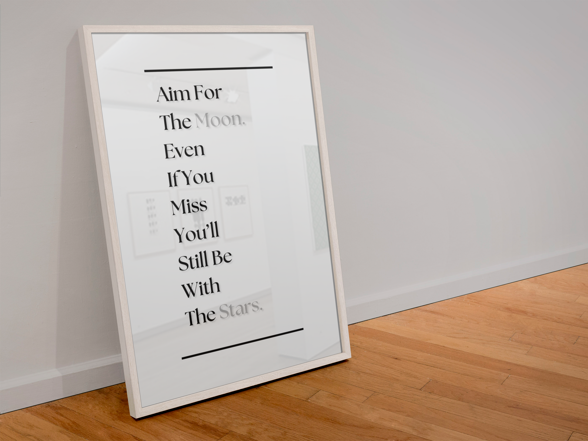 A framed typographic wall art print leaning against a light gray wall on a wood floor. The print features black text reading, 'Aim For The Moon. Even If You Miss You'll Still Be With The Stars'.