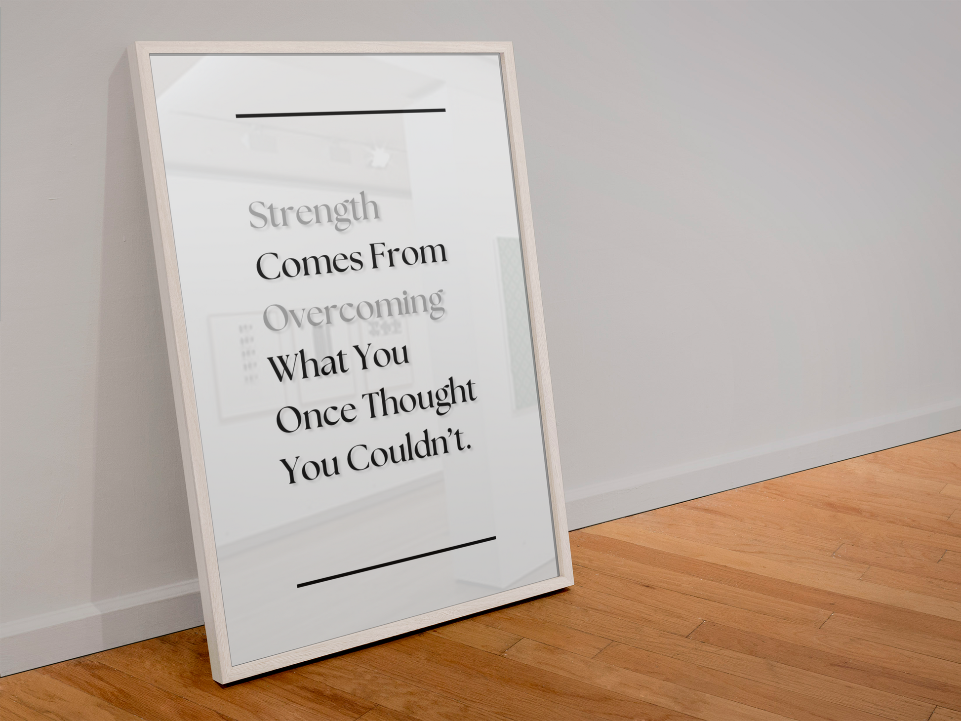 A framed motivational quote print leaning against a light gray wall on a wooden floor. The print features a white background with black and gray text reading 'Strength Comes From Overcoming What You Once Thought You Couldn't.'