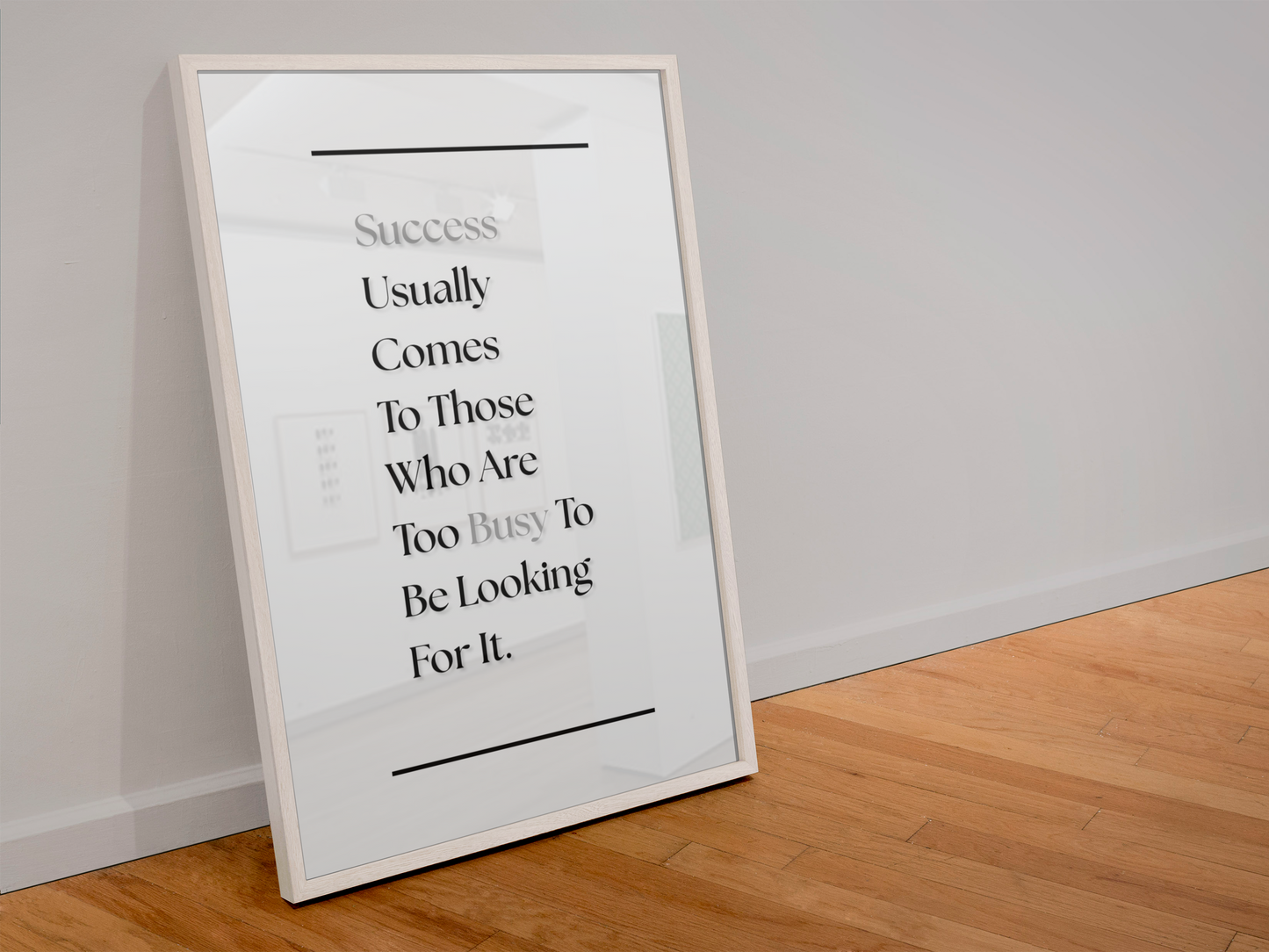A framed motivational art print leaning against a white wall on a light wood floor. The print features the quote 'Success Usually Comes To Those Who Are Too Busy To Be Looking For It'.