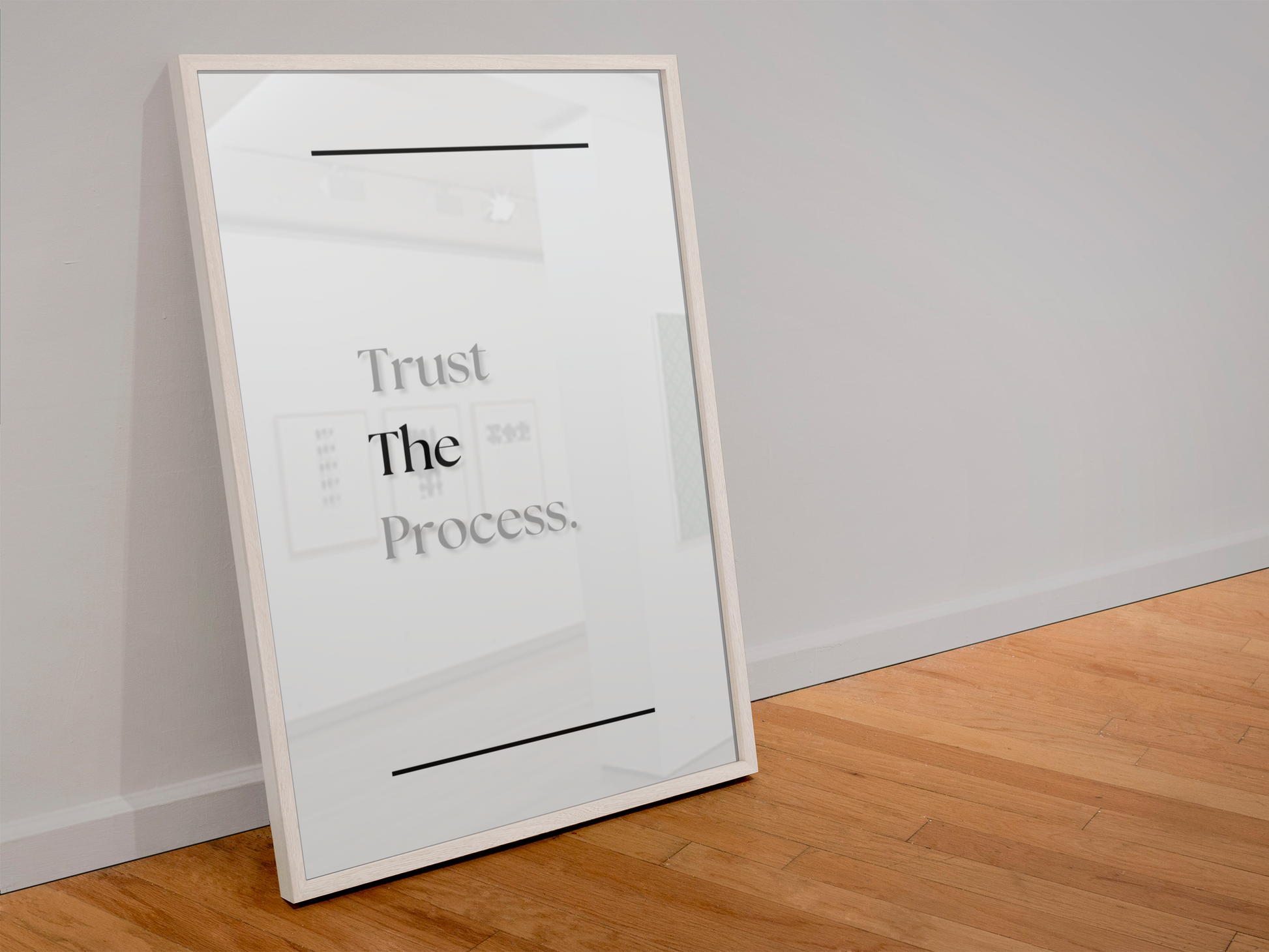 A framed minimalist art print leaning against a white wall on a light wood floor. The print features the text 'Trust The Process.'