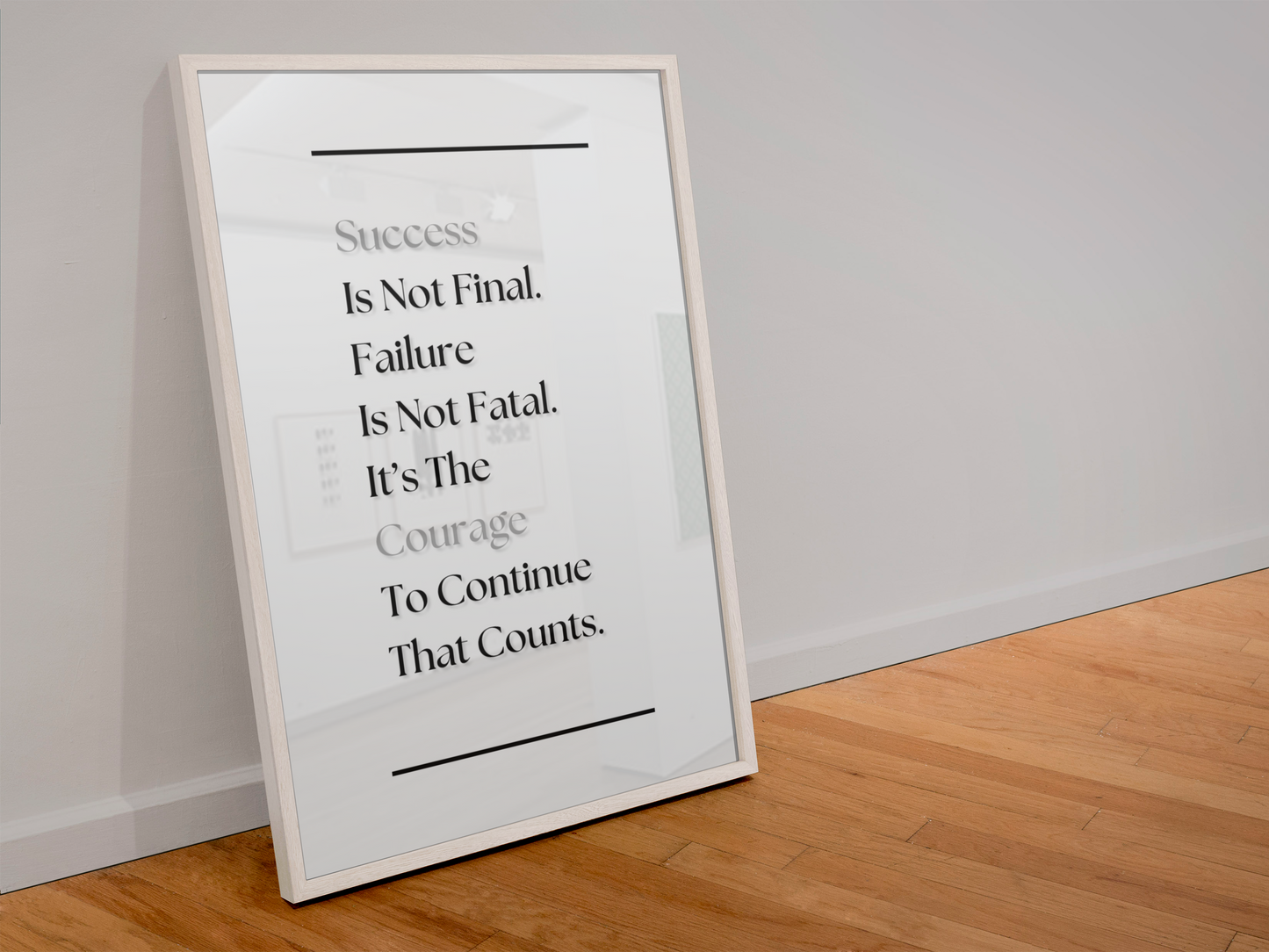 A framed motivational quote print leaning against a white wall on a light wood floor. The print features a white background with black and gray text reading, 'Success Is Not Final. Failure Is Not Fatal. It's The Courage To Continue That Counts'.